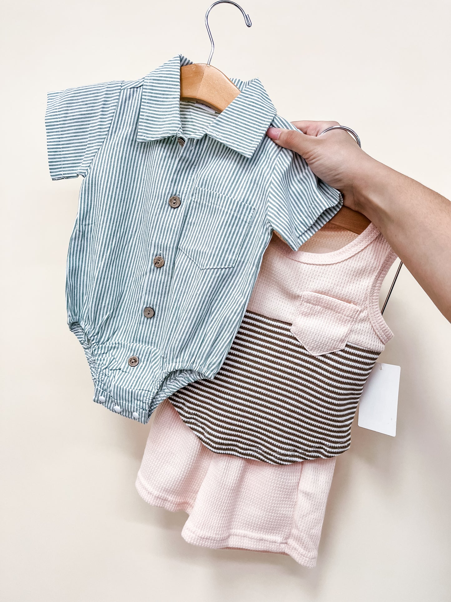 A hand holds Tiny Tot Bundles’ Bundle Of The Day - Boys: two baby outfits on wooden hangers—a blue and white striped short-sleeve romper with buttons, and a sleeveless pink dress with a brown and cream striped bodice and small pocket.