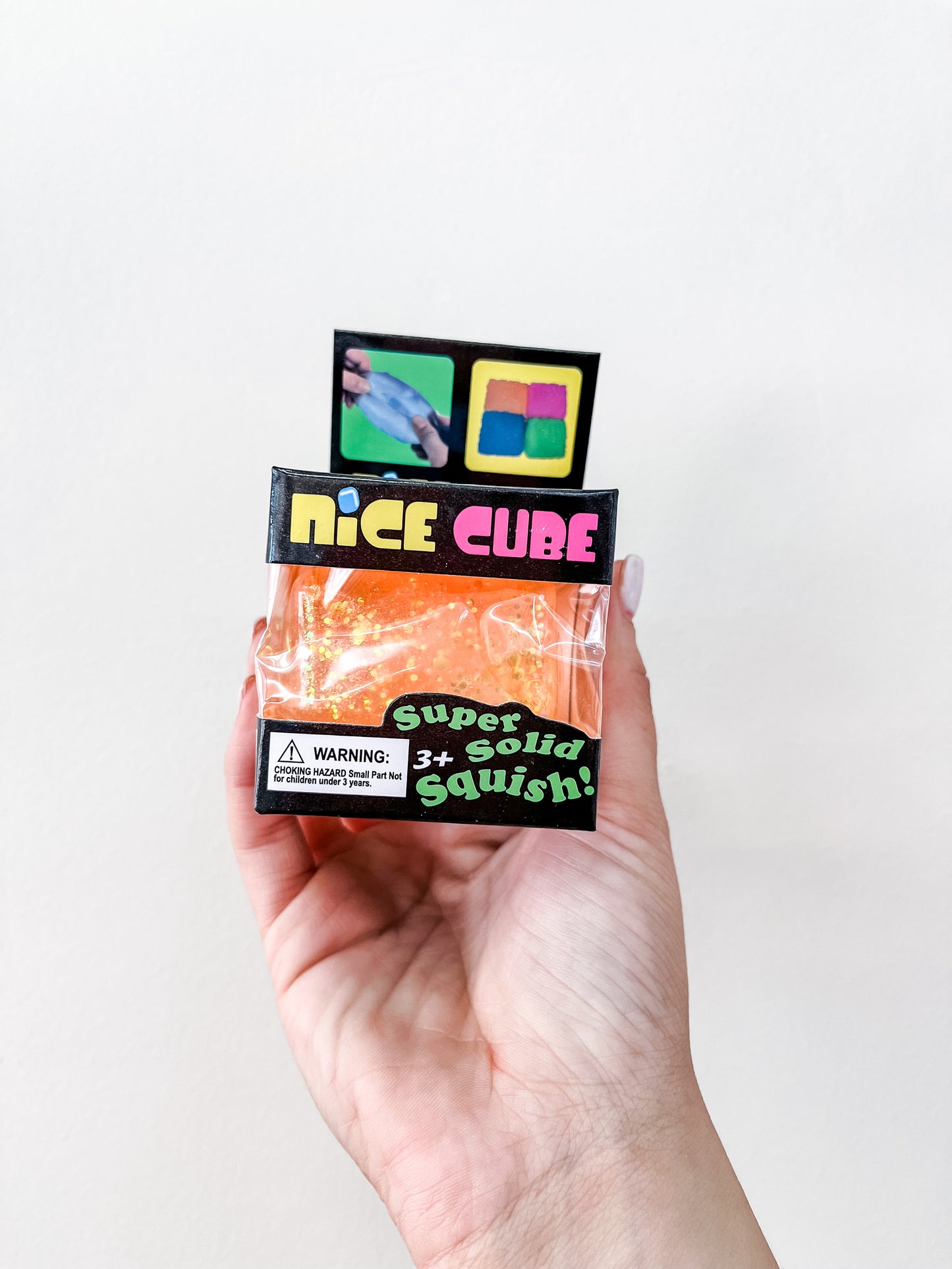 A hand holds a package labeled Prime Opticals Glitter Squishy Cube containing an orange, glittery squishy toy. The packaging says Super Solid Squish! with colorful cube images and a small warning label visible.