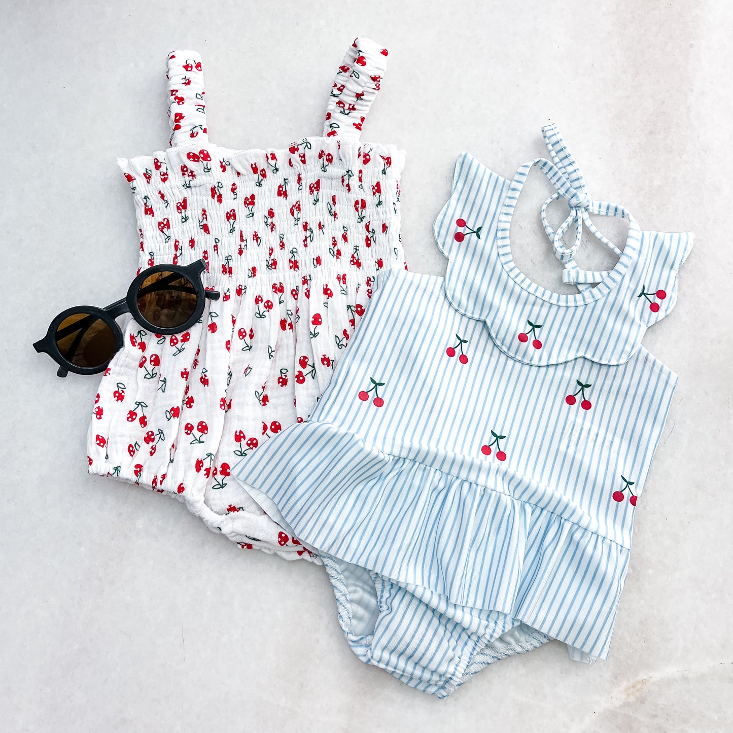 Two swimsuits from Tiny Tot Bundles' "Bundle Of The Day - Girls" are displayed: one white with red cherries and straps, the other light blue with stripes, ruffled collar, and skirt. Black round sunglasses lie beside them.