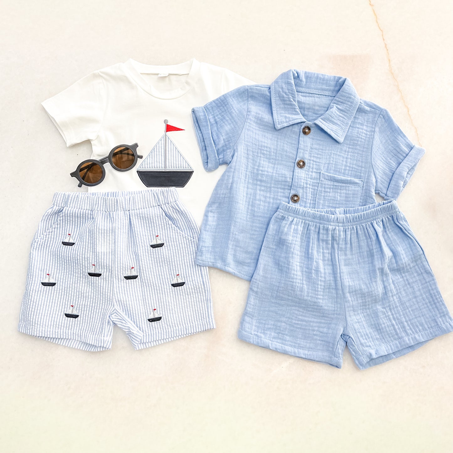 Flat lay of Bundle Of The Day - Boys by tiny: a white sailboat graphic tee, blue striped boat shorts, light blue button-up shirt with matching shorts, and round sunglasses.