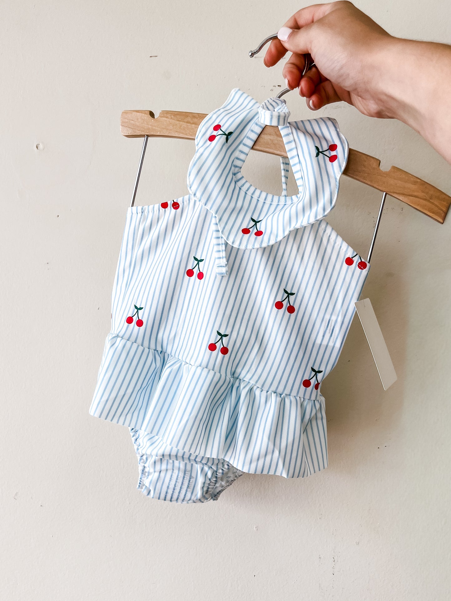 A hand holds a hanger with the Tiny Tot Bundles Cherry Stripe Swimsuit, a baby girls' blue and white striped romper featuring red cherry prints, ruffled collar, and ruffled bottom, displayed against a plain light-colored wall.