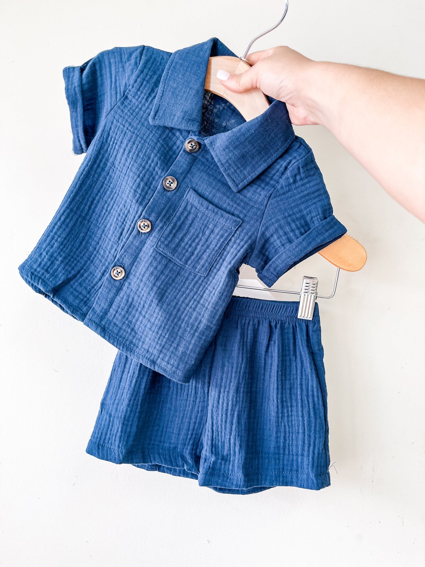 A hand holds a hanger with the Navy Linen Set by Tiny Tot Bundles—featuring a blue short-sleeve button-up shirt and matching shorts in soft, textured fabric—displayed against a plain white background.