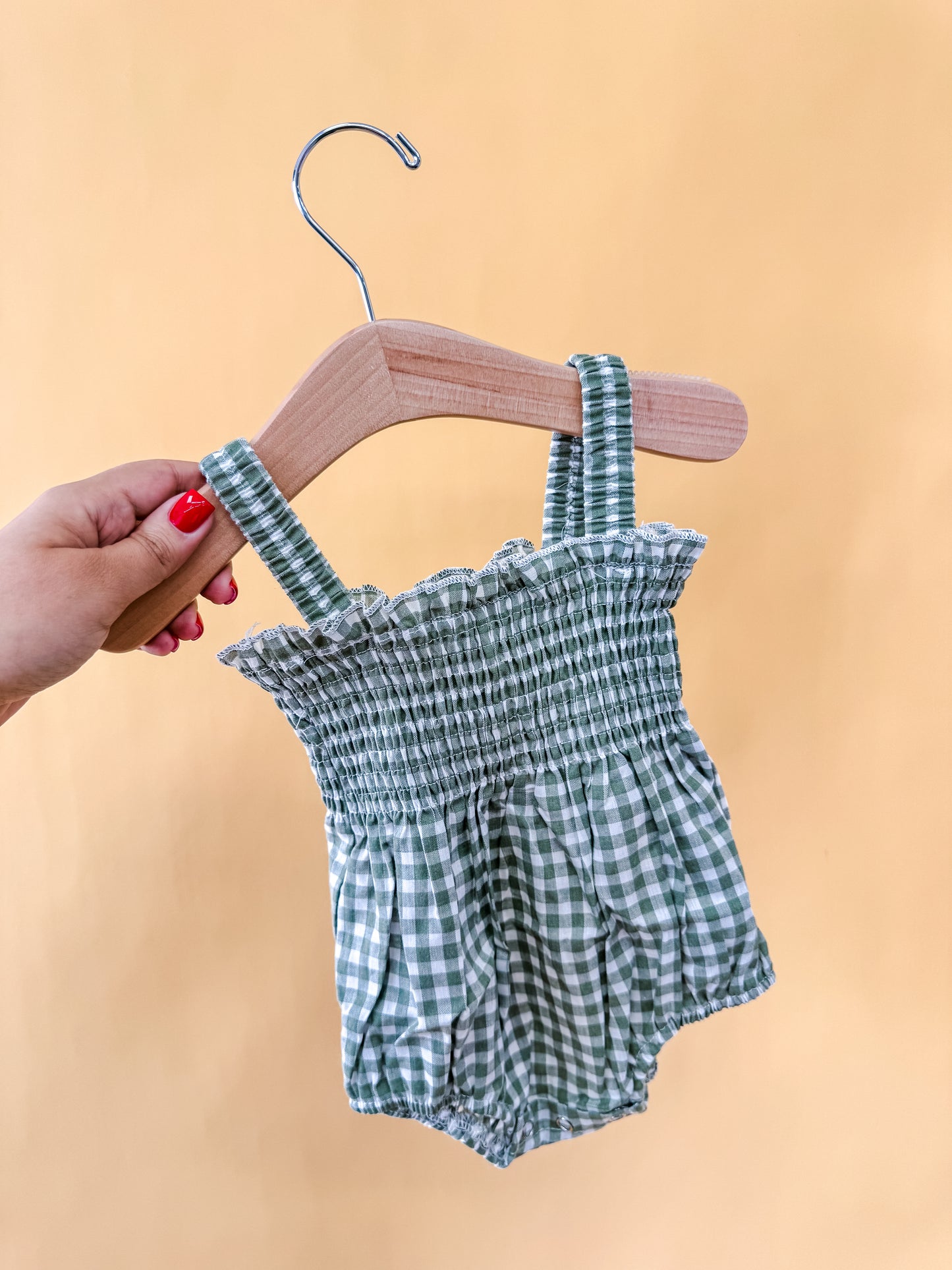 A hand with red nail polish holds a wooden hanger displaying the Meadow Mist Romper by Tiny Tot Bundles, a green and white checkered baby romper with ruffled trim and suspenders, set against a plain light yellow background.