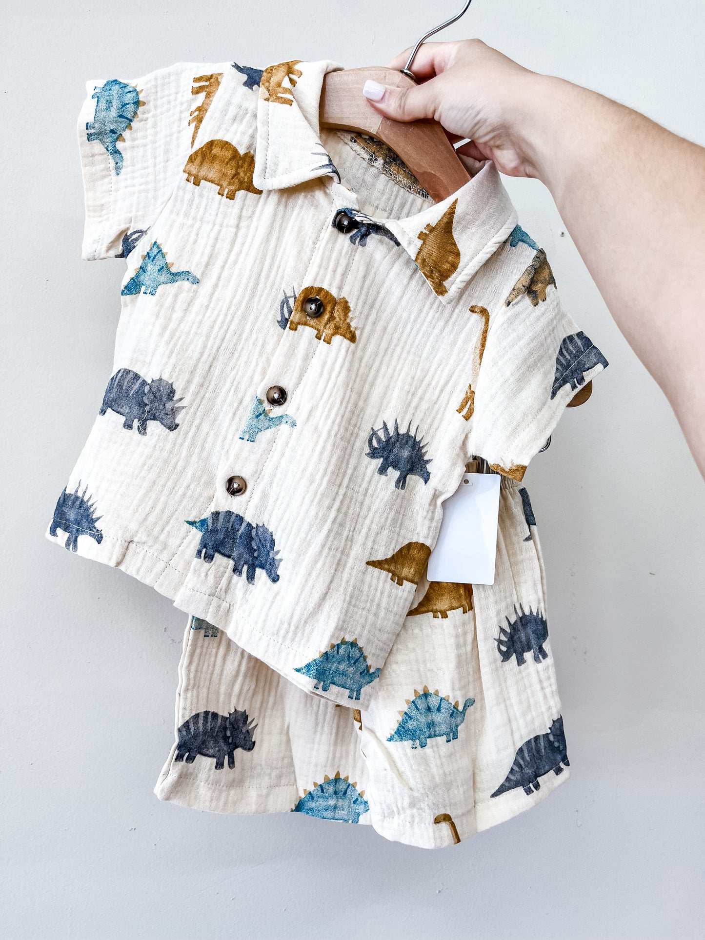 A hand holds up the Dino Linen Collard Set by Tiny Tot Bundles, a matching short-sleeve shirt and shorts for kids, made of cream-colored fabric with vibrant blue, mustard yellow, and gray dinosaur prints.