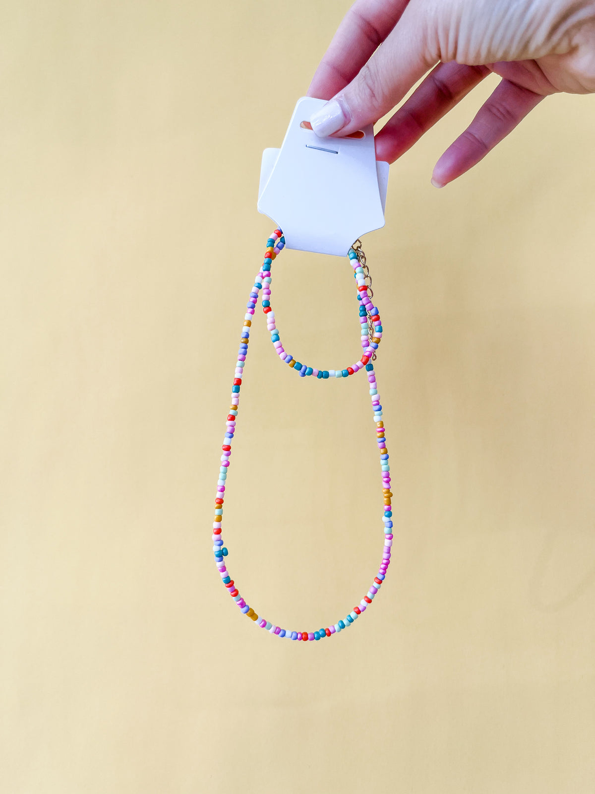 A hand with light pink nail polish holds a Tiny Tot Bundles Summer Beaded Bracelet Set on a display card, set against a plain yellow background.