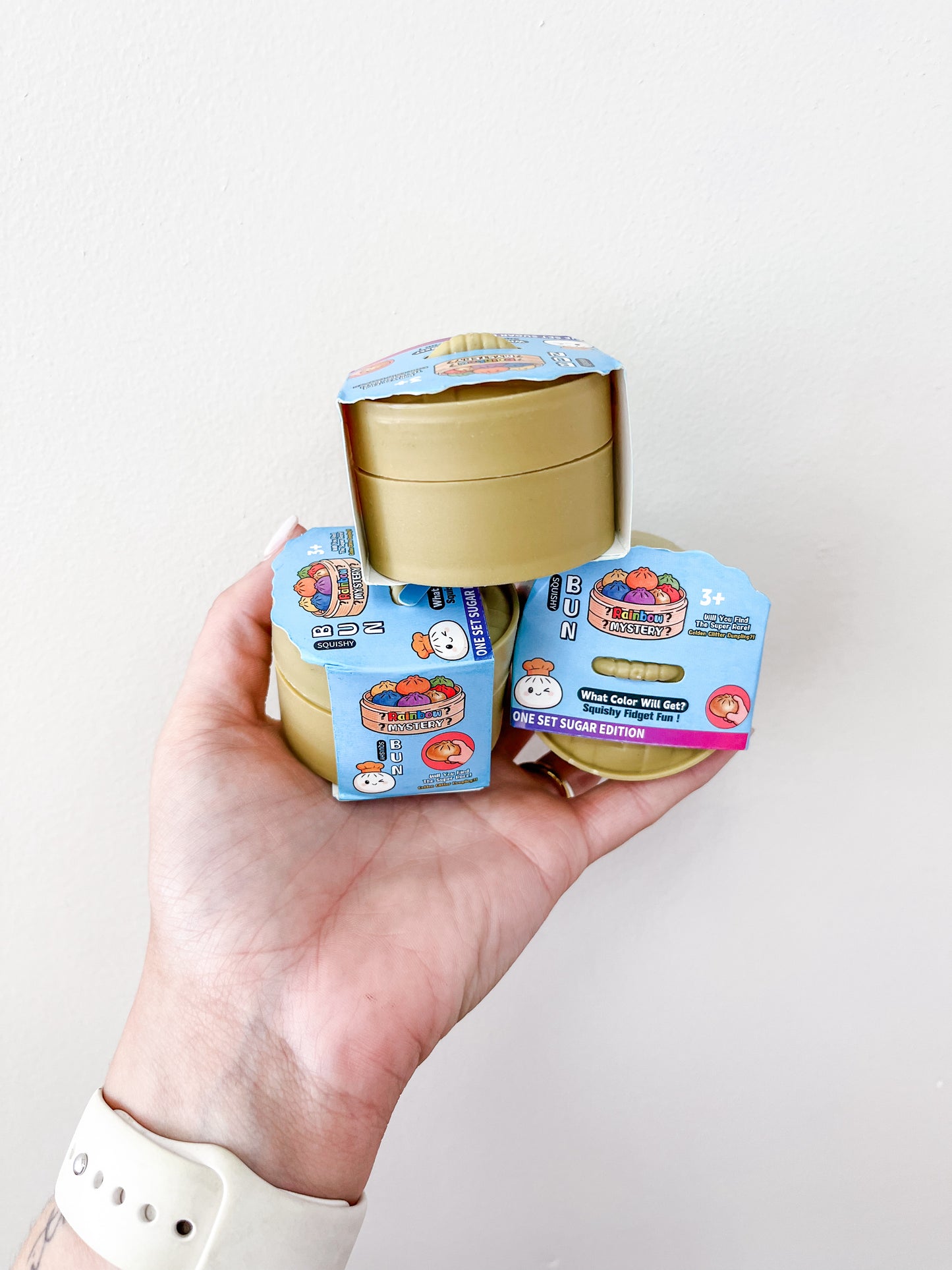 A hand holds three MINI Gel Glitter Dumpling containers from Prime Opticals, stacked in the palm. The round packages have blue and gold designs, featuring colorful graphics and text, set against a plain background.