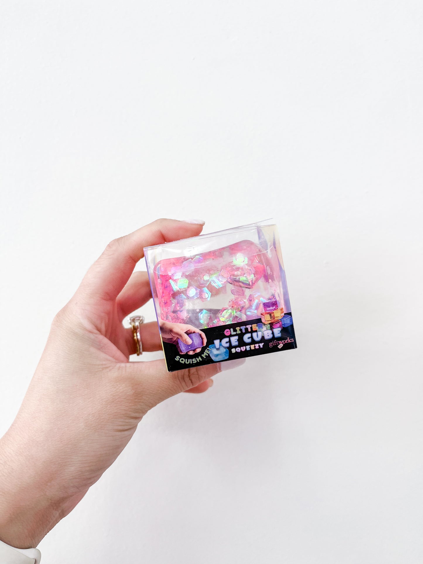 A hand holds a small clear box with a pink, glittery, iridescent squishy toy labeled "Confetti Glitter Ice Cubes" by Prime Opticals against a plain white background.