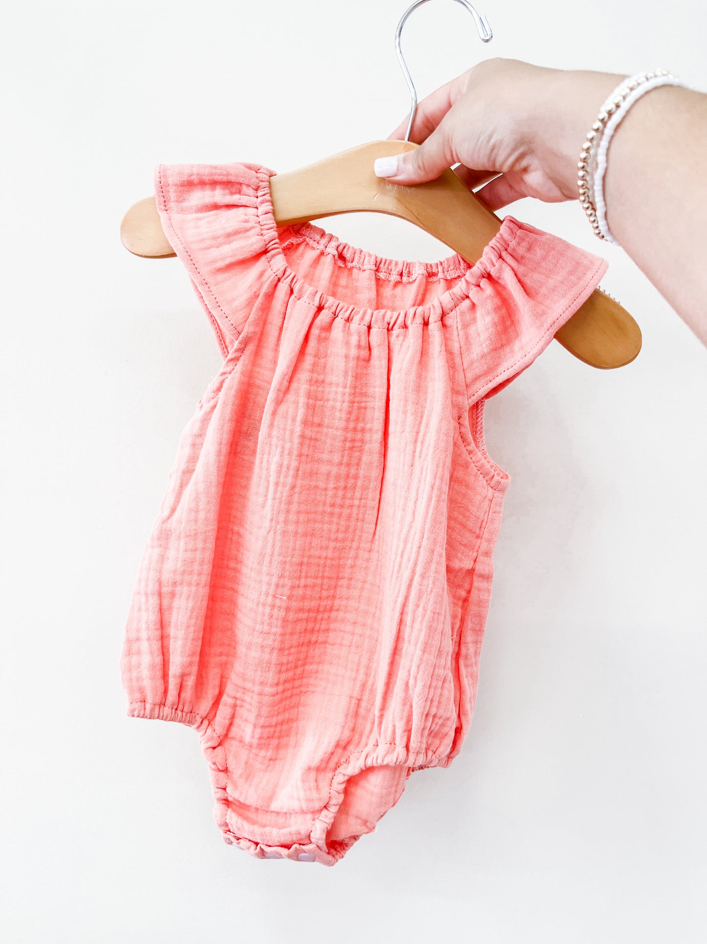 A hand holds the Tiny Tot Bundles Pink Summer Linen Romper, featuring ruffled sleeves and gathered edges, displayed on a wooden hanger against a plain white background.
