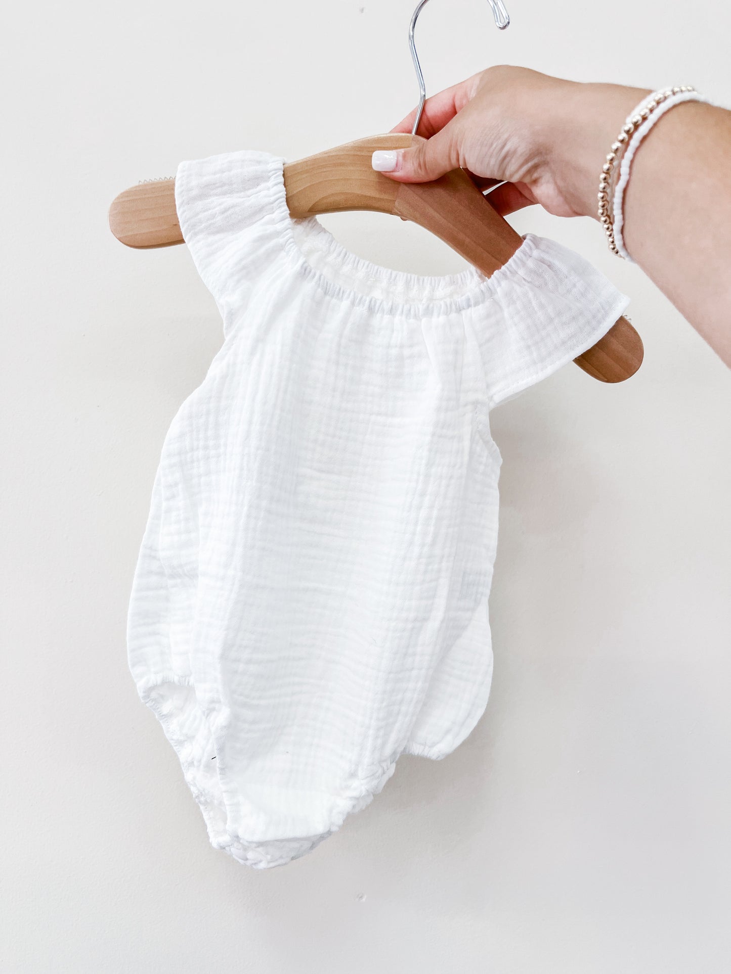 A hand holds a Tiny Tot Bundles White Summer Linen Romper, made of lightweight, textured fabric, displayed on a wooden hanger against a plain white background.