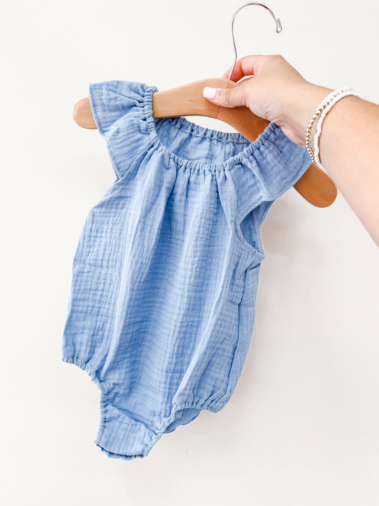 A hand holds a wooden hanger with the Tiny Tot Bundles Blue Summer Linen Romper, featuring light blue ruffles, shown against a plain white background.