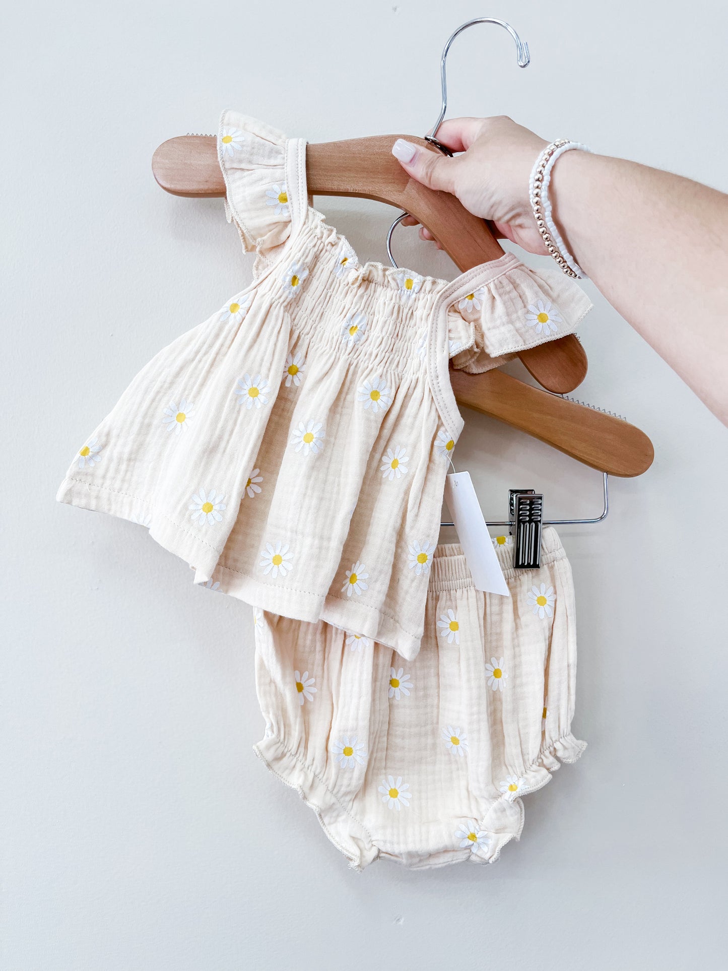 A hand holds the Tiny Tot Bundles Beige Daisy Set—matching baby top with ruffled sleeves and daisy prints, and coordinating bloomers—against a plain light background.