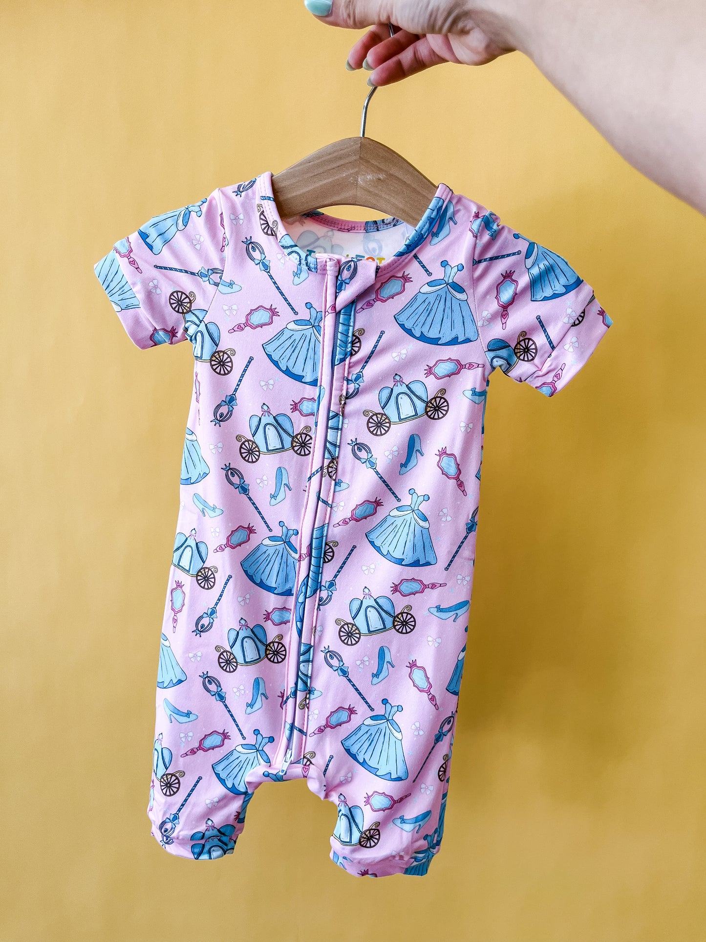 A hand holds a wooden hanger displaying the Tiny Tot Bundles Royal Ball Short Zippy, a pink baby romper adorned with blue dresses, shoes, carriages, and tiaras, set against a yellow background.
