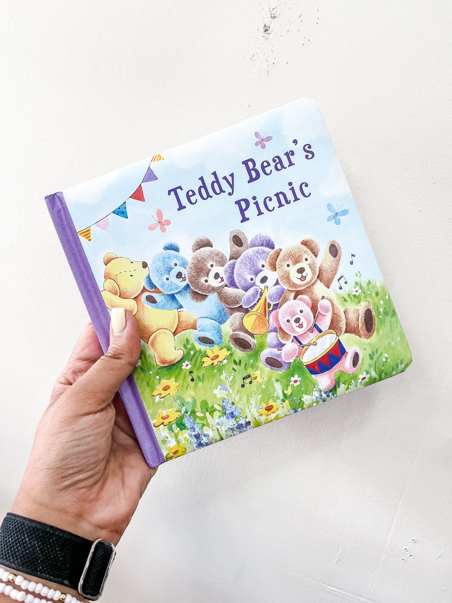 A hand holds the Tiny Tot Bundles Teddy Bear's Picnic Book, featuring illustrated teddy bears sitting on grass with flowers, bunting, and musical notes.