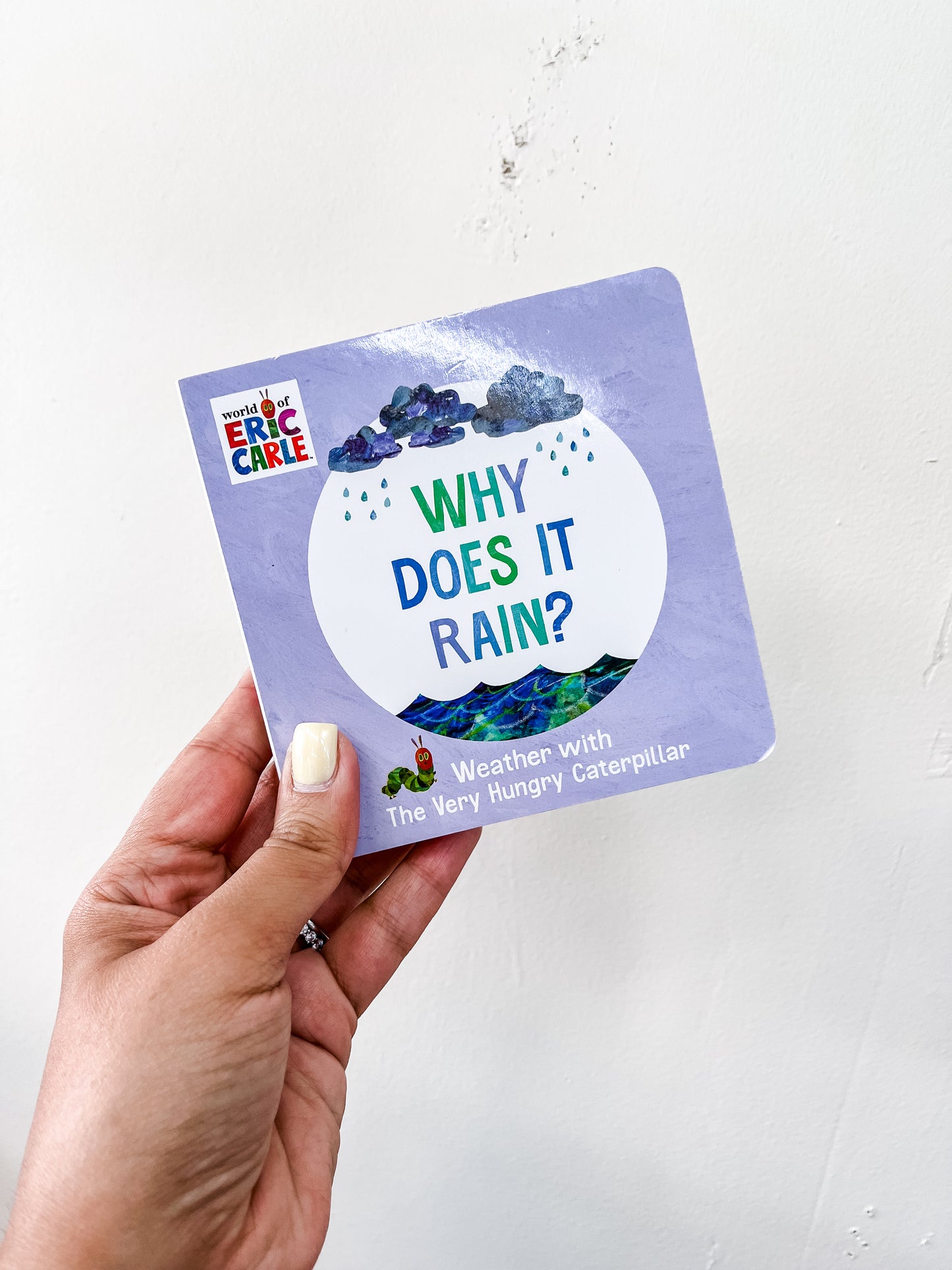 A hand with light-colored nail polish holds the Tiny Tot Bundles Why Does It Rain Book against a plain white background.