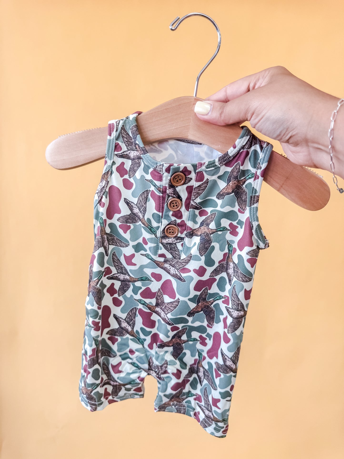 A hand holds a hanger displaying the Tiny Tot Bundles Mallard Marsh Tank Romper, a sleeveless baby outfit adorned with a green, brown, and tan bird and camouflage pattern and detailed with two wooden buttons, set against a yellow background.
