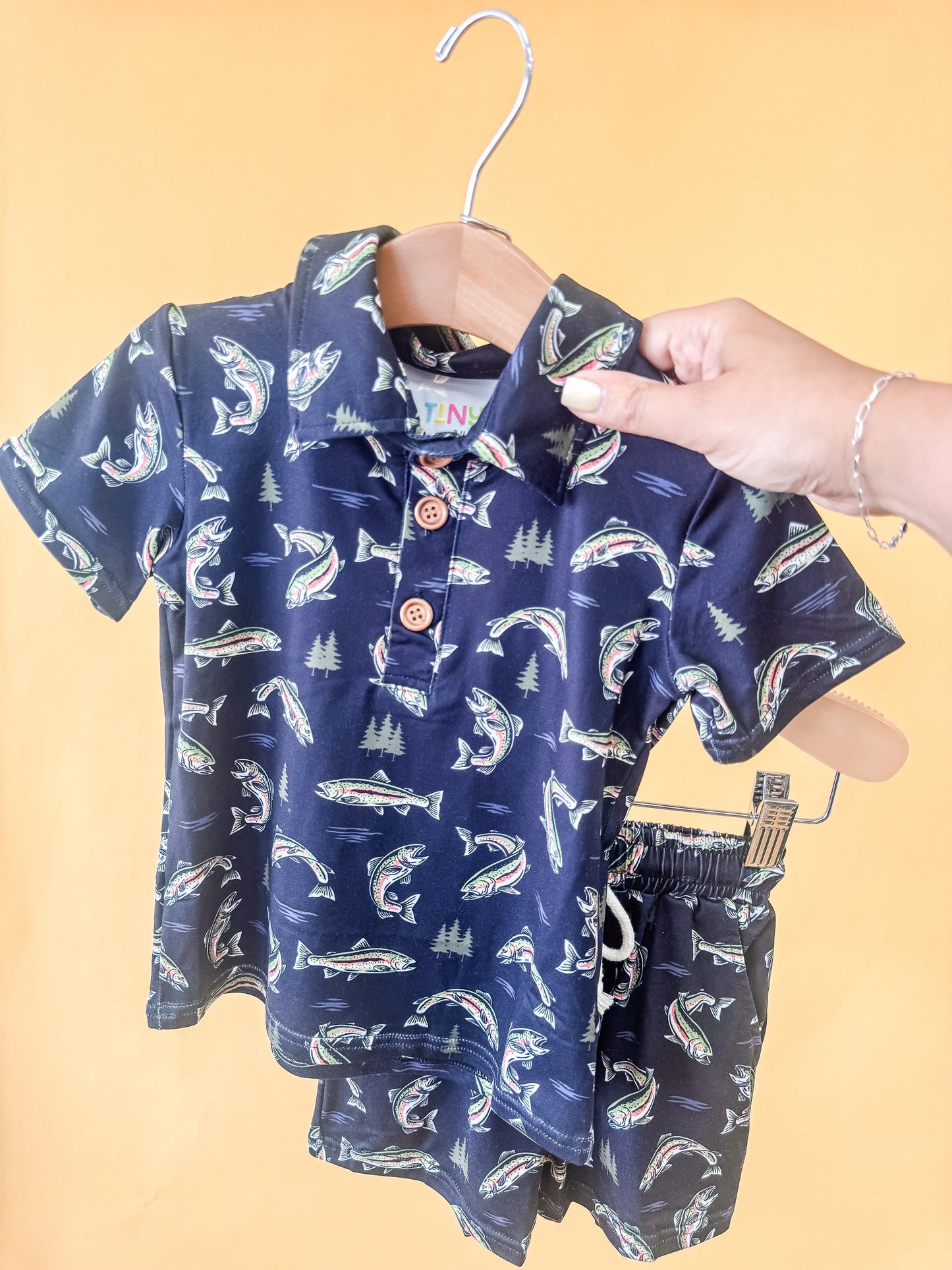 The "Reel Adventure Collared Set" by Tiny Tot Bundles, featuring navy blue toddler shirt and shorts adorned with fish and tree patterns, hangs on a hanger against a yellow background. A hand holds the collar to highlight its detail.