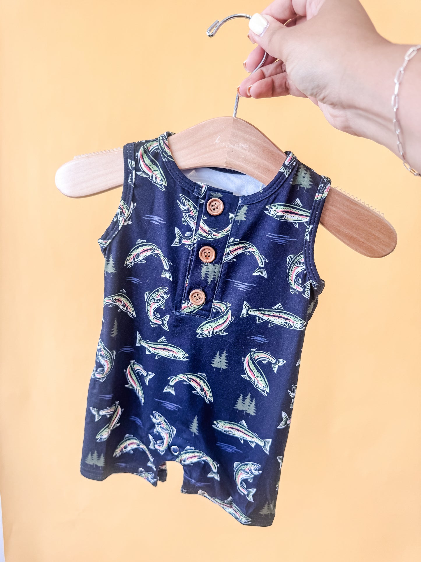 A hand holds Tiny Tot Bundles' Reel Adventure Tank Romper, a sleeveless baby outfit with fish and tree patterns on a navy background, displayed against a yellow backdrop.