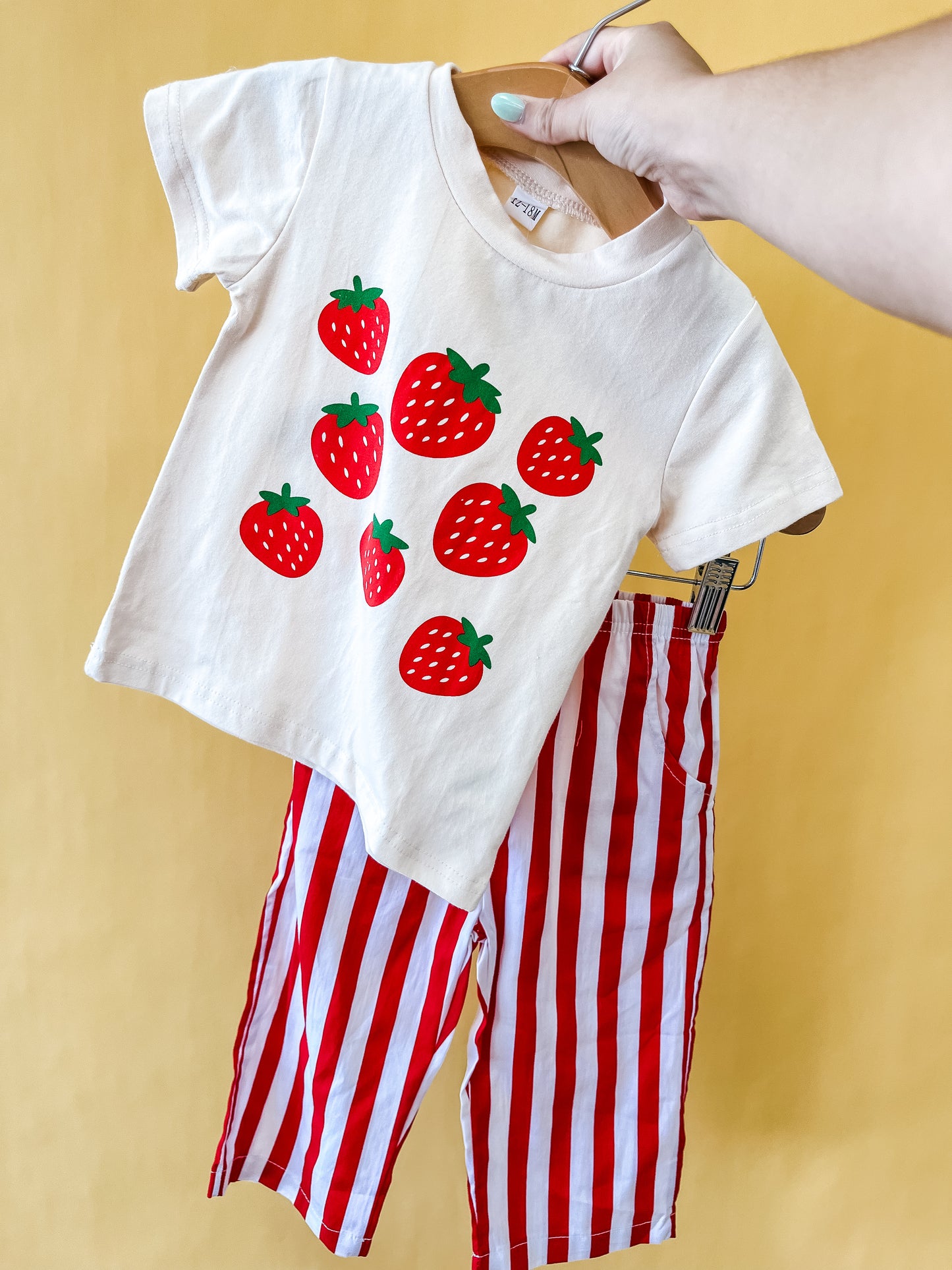 A hand holds the Tiny Tot Bundles Strawberry Patch Set, featuring a children's white T-shirt adorned with red strawberries and matching red-and-white vertically striped pants, set against a yellow background.