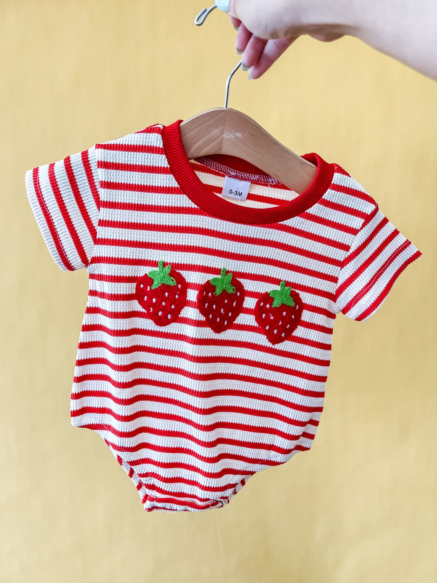 A hand holds the Tiny Tot Bundles "Sweet as Strawberry Bubble" baby romper, featuring red and white stripes and three strawberry appliqués on the front, displayed on a wooden hanger against a yellow background.