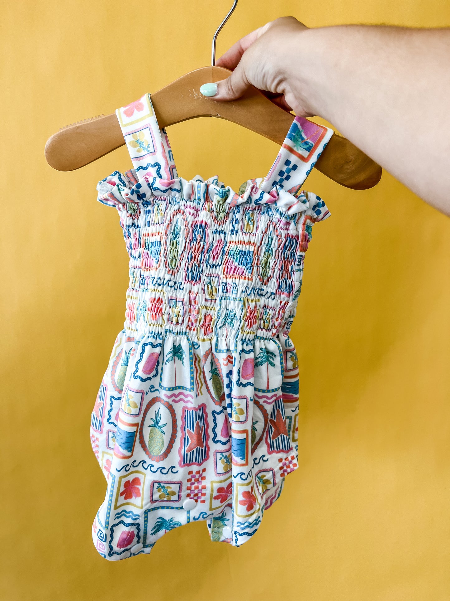 A hand holds a wooden hanger displaying the Tiny Tot Bundles Sunshine Souvenir Smocked Romper—a colorful, patterned baby romper with ruffled, smocked fabric and wide straps—against a bright yellow background.