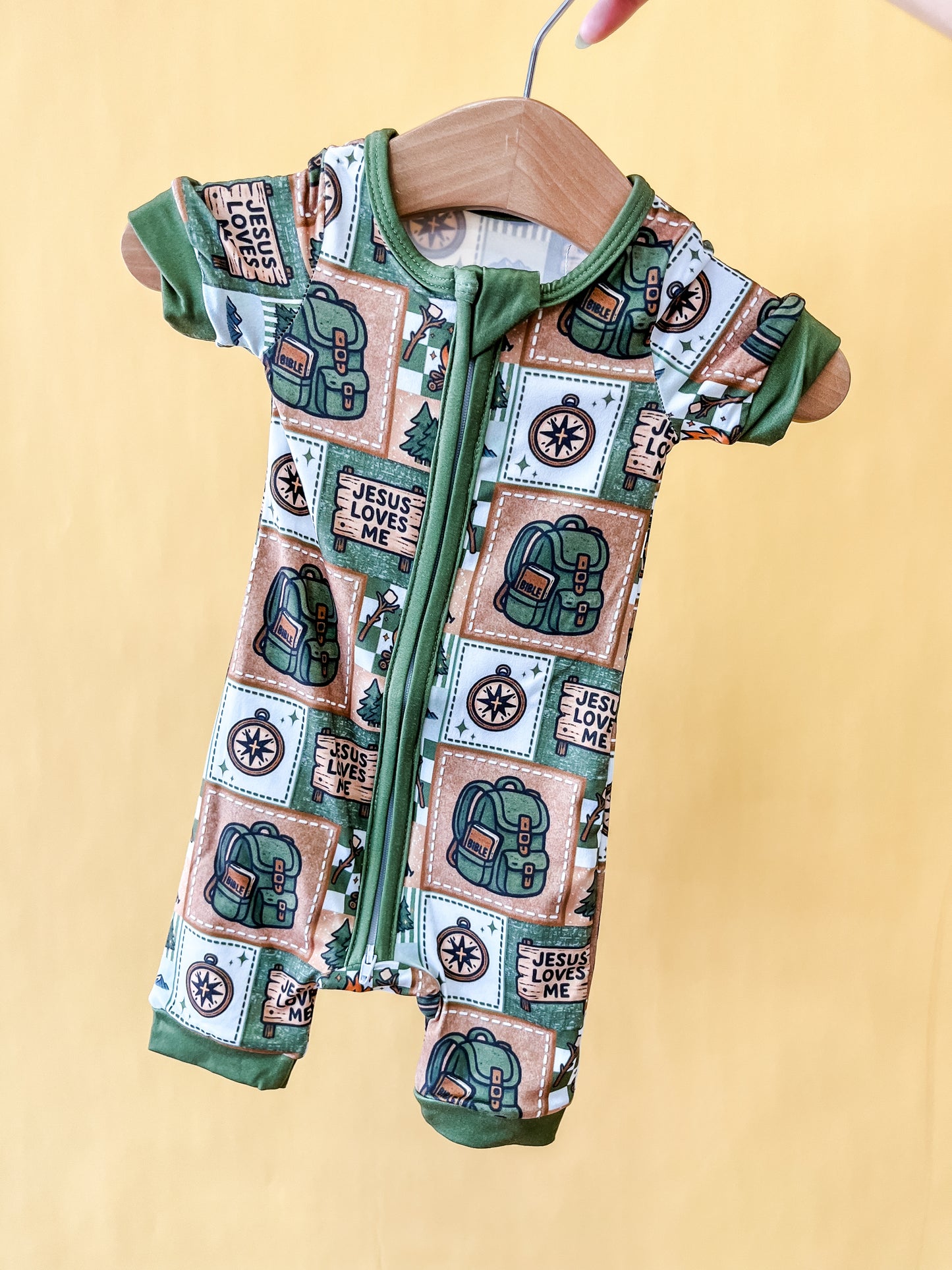 The Camper's Faith Short Zippy by Tiny Tot Bundles is a short-sleeved baby romper featuring green trim, a front zipper, and playful prints of Baby Yoda, stars, and the phrase "Jesus Loves Me" on a beige background, displayed on a wooden hanger.
