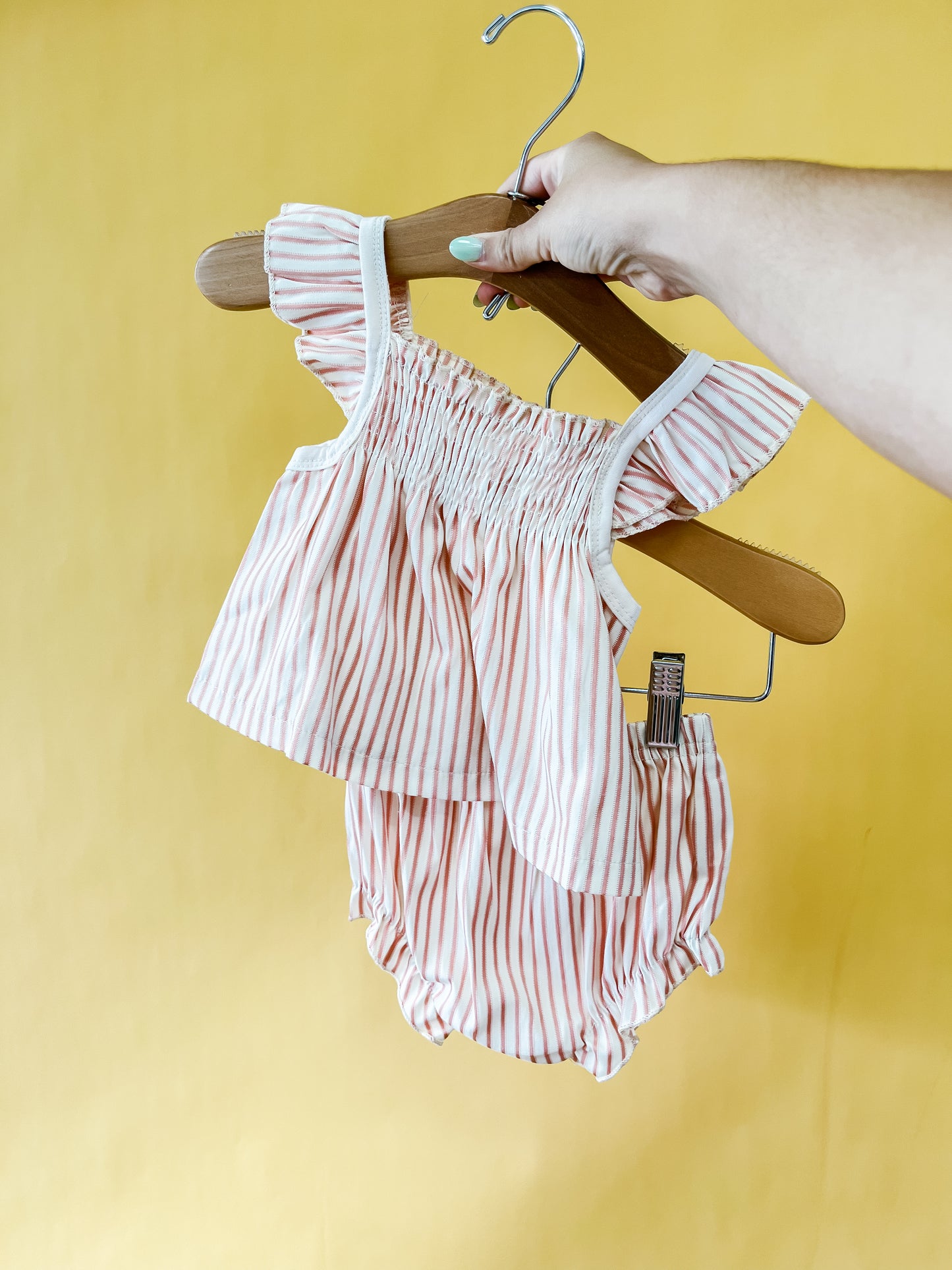 A hand holds a hanger with the Tiny Tot Bundles Rosy Picnic Set, a pink-and-white striped baby outfit featuring a ruffled top and matching bloomers, against a yellow background.