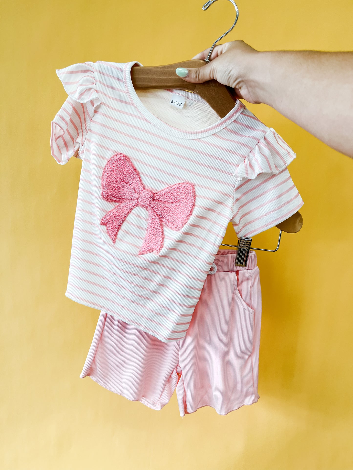 A hand holds the Bow Stripe Set by Tiny Tot Bundles, featuring a child’s pink and white striped top with a large pink bow and matching pink shorts, set against a yellow background.