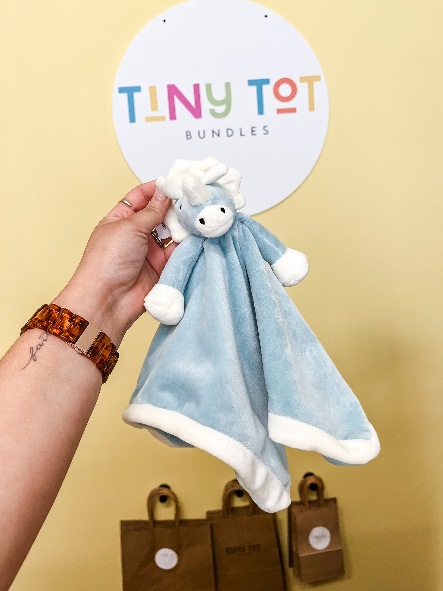 A hand holds the Snuggle Plush by Tiny Tot Bundles, a soft blue-and-white toy with a lamb’s head, in front of a round Tiny Tot Bundles sign and brown paper bags in the background.