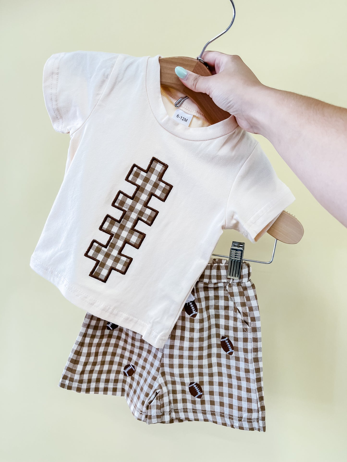 A hand holds up the Tiny Tot Bundles Kickoff Cutie Short Set, featuring a cream t-shirt with a football lace design and brown checkered shorts adorned with small football prints, against a pale yellow background.