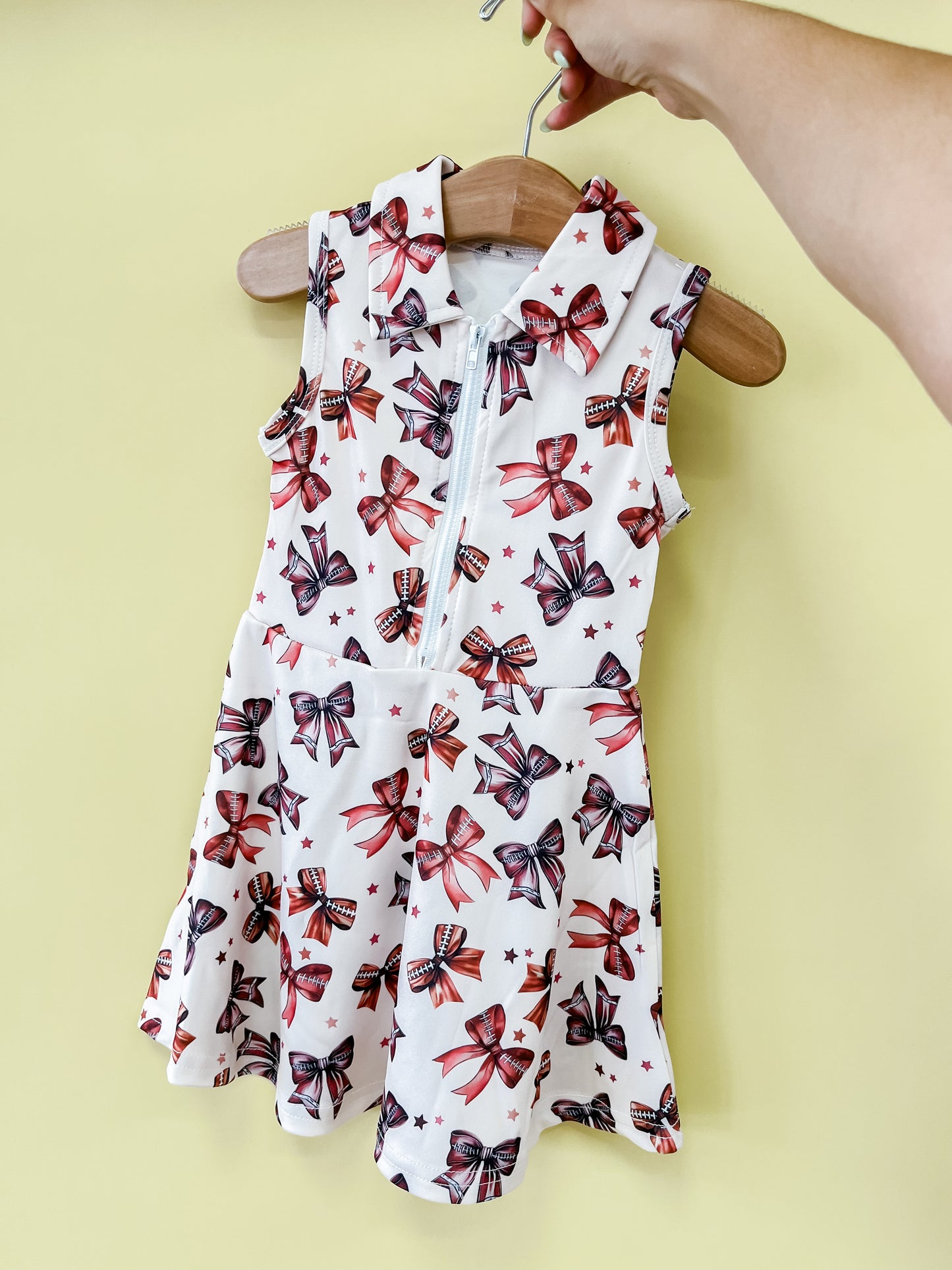The Football & Bows Dress by Tiny Tot Bundles, a sleeveless white collared dress featuring red, blue, and black bow patterns, is displayed on a wooden hanger against a pale yellow background.