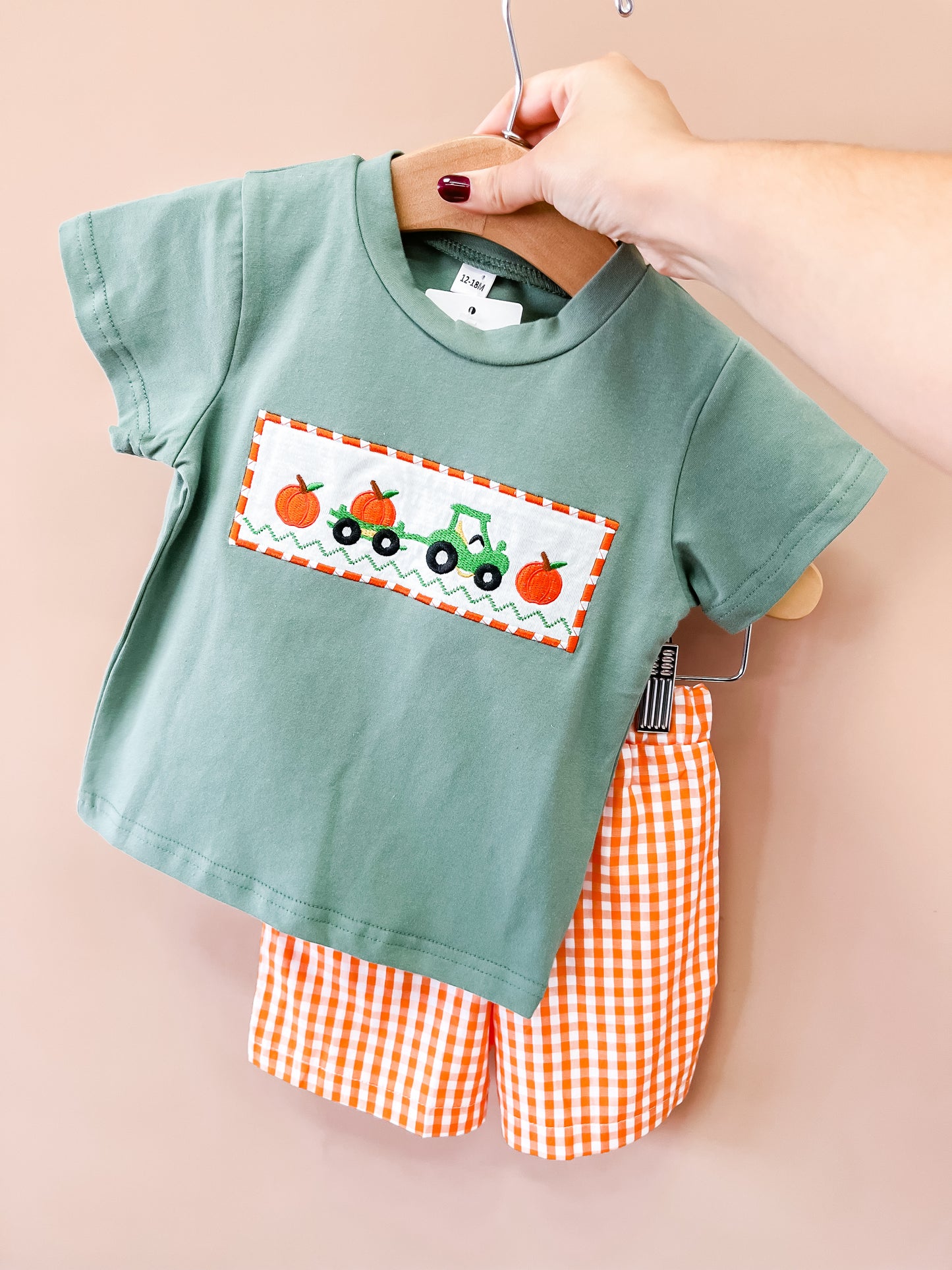 A person with painted nails holds the Tiny Tot Bundles Pumpkin Patch Tractor Set on a hanger, featuring a green toddler t-shirt with a tractor and pumpkin design, paired with orange and white checkered shorts.