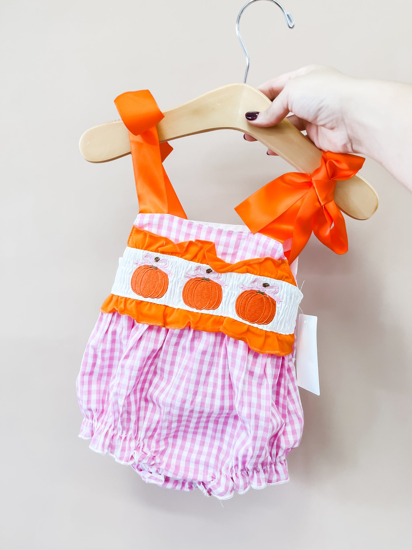 A hand holds the Little Miss Pumpkin Romper by Tiny Tot Bundles—a pink gingham baby romper featuring orange ribbons and embroidered pumpkins on the front—displayed on a wooden hanger against a light beige background.
