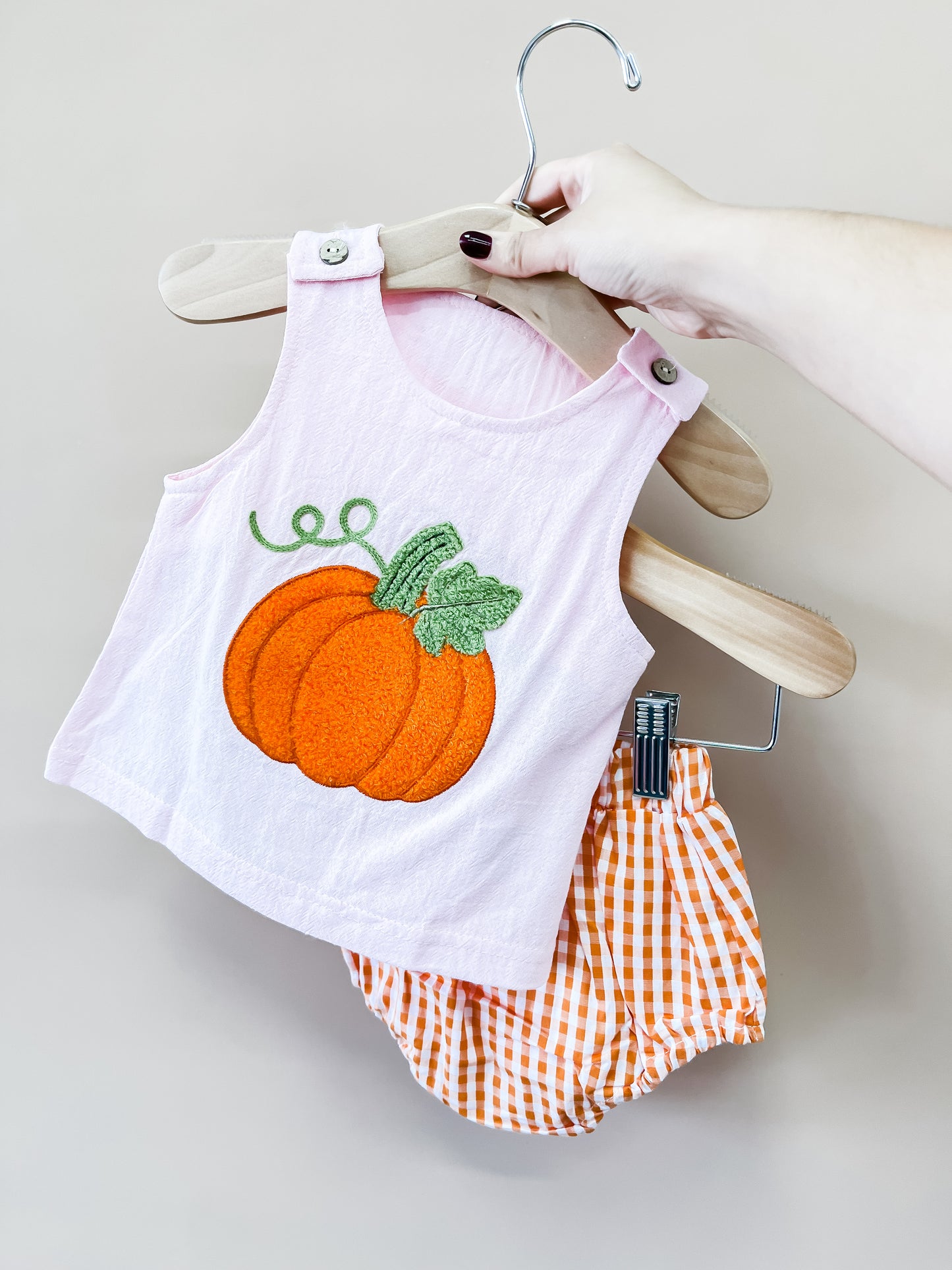 A hand holds a hanger with Tiny Tot Bundles’ Blushing Pumpkin Set—a pink toddler tank top adorned with a pumpkin patch and matching orange gingham shorts.