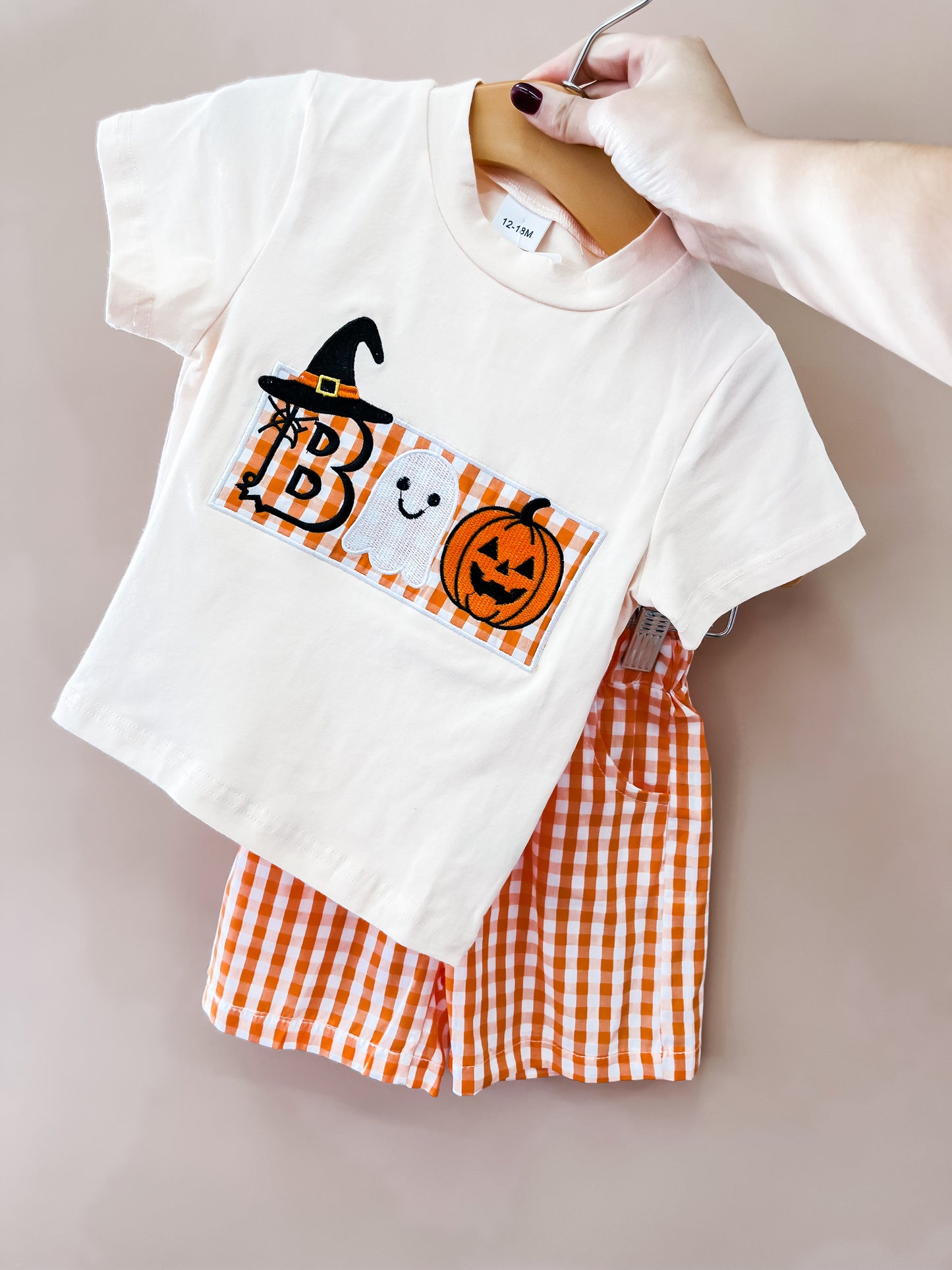 A hand holds the Tiny Tot Bundles Boo Gingham Halloween Set, which includes a white shirt decorated with Halloween-themed “BOO” letters, a witch hat, ghost, and pumpkin, paired with orange gingham shorts.