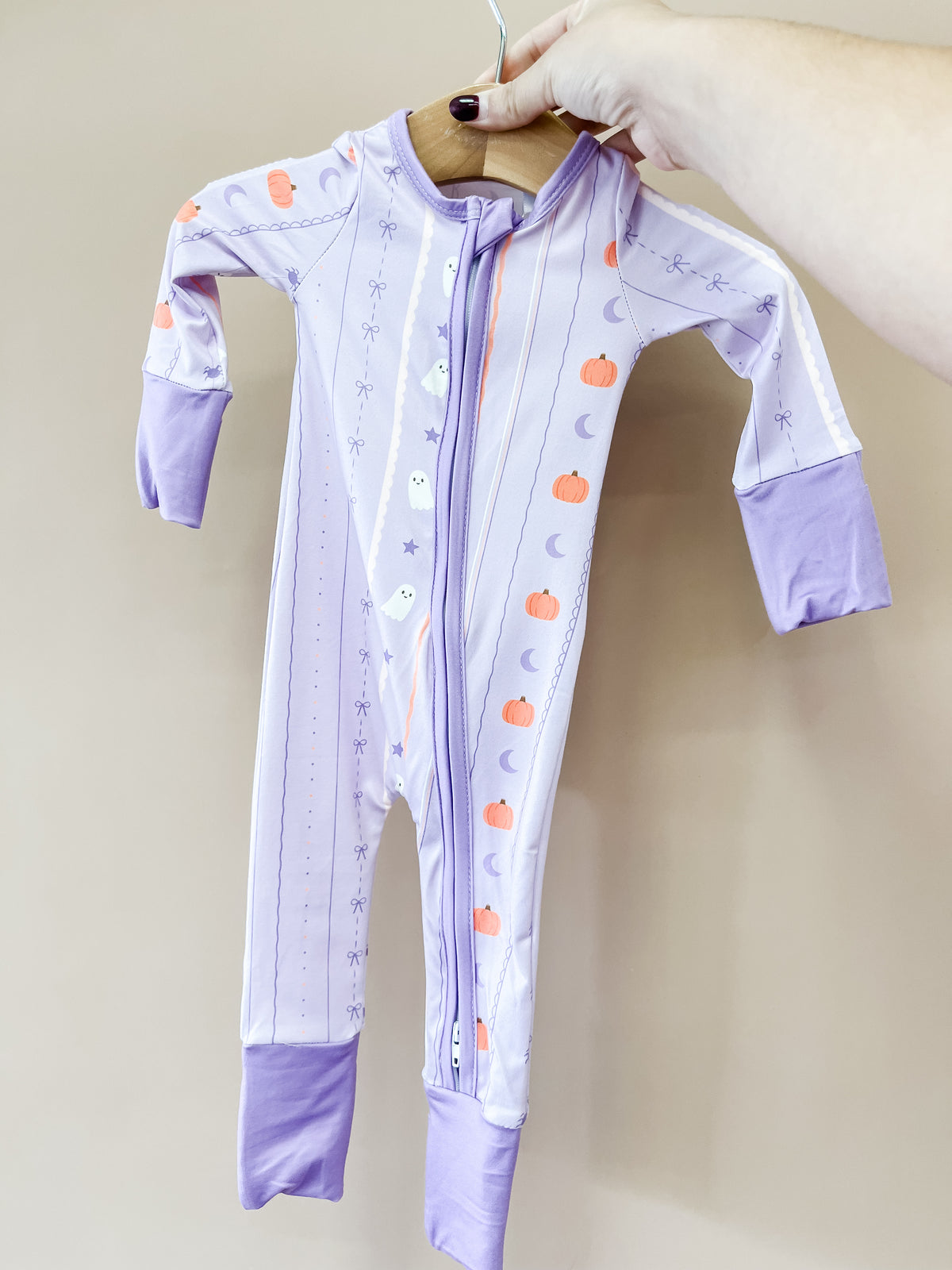 A hand holds up the Lavender Haunt Zippy by Tiny Tot Bundles, a lavender baby onesie with purple cuffs, a front zipper, and decorated with pumpkins, ghosts, moons, and stars.