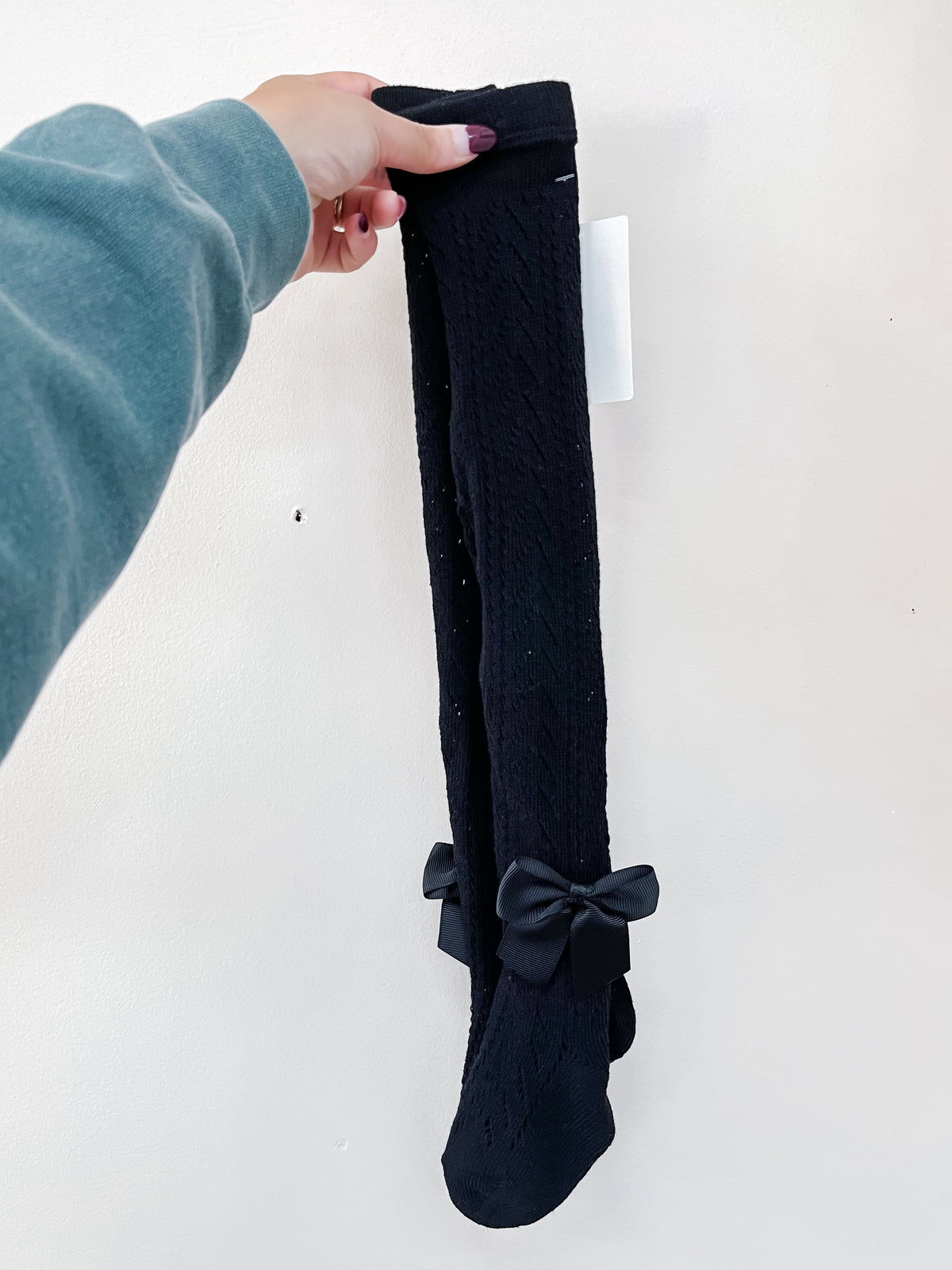 A hand holds up a pair of black Sweet Bow Detail Knit Socks featuring a textured cable-knit pattern and a small ribbon bow at the ankle.