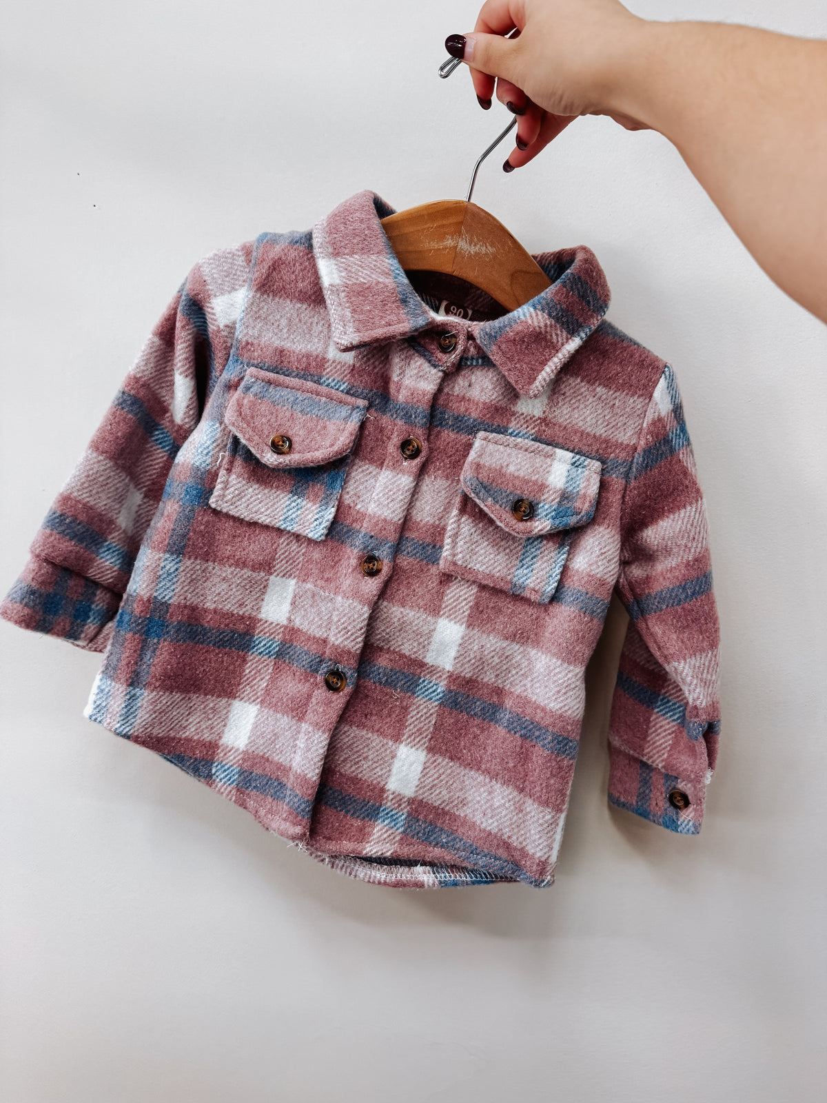 A hand holds a Blush Pink Flannel in blush pink, blue, and white plaid on a wooden hanger.
