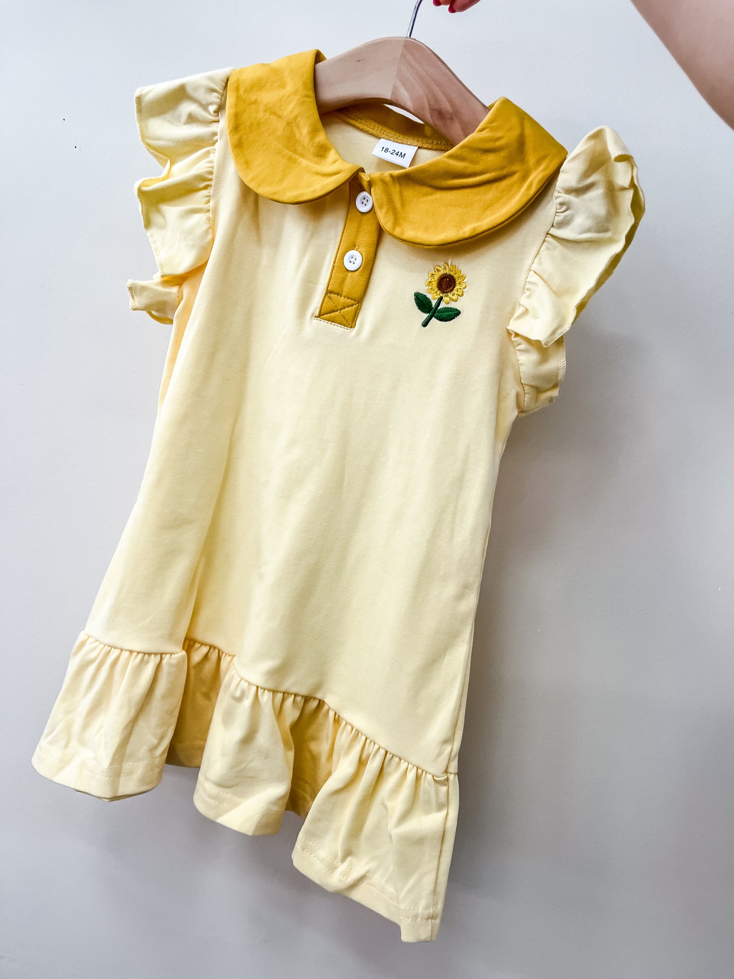 The Sunflower Ruffles Dress by Tiny Tot Bundles is a light yellow girls’ dress with ruffled sleeves, a yellow collar, front buttons, and sunflower embroidery on the chest, shown hanging on a wooden hanger against a simple background.