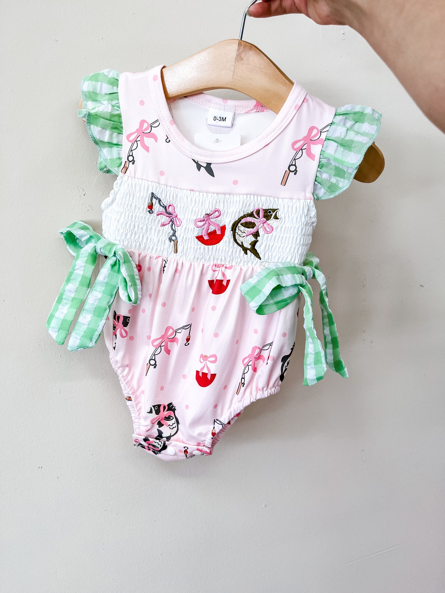The Pretty Pink Fishing Romper by Tiny Tot Bundles features green gingham ruffle sleeves, side bows, and an adorable print of cats, umbrellas, and pink bows. Displayed on a wooden hanger and held by hand.