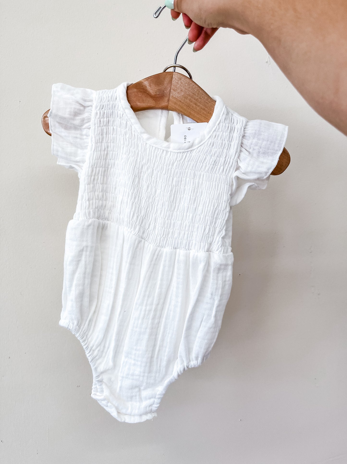 A hand holds a wooden hanger displaying the White Timeless Romper by Tiny Tot Bundles, which features short, ruffled sleeves and a textured top, set against a plain light-colored wall.