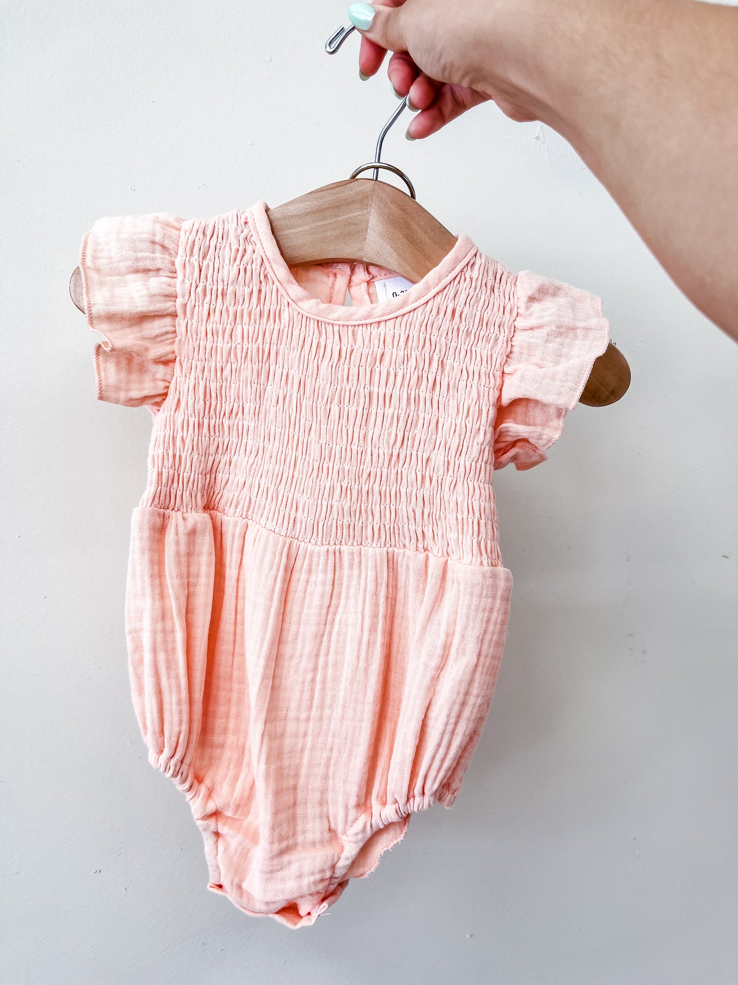 A hand holds a wooden hanger with the Peach Timeless Romper by Tiny Tot Bundles, featuring short, ruffled sleeves and a smocked bodice, set against a plain white background.