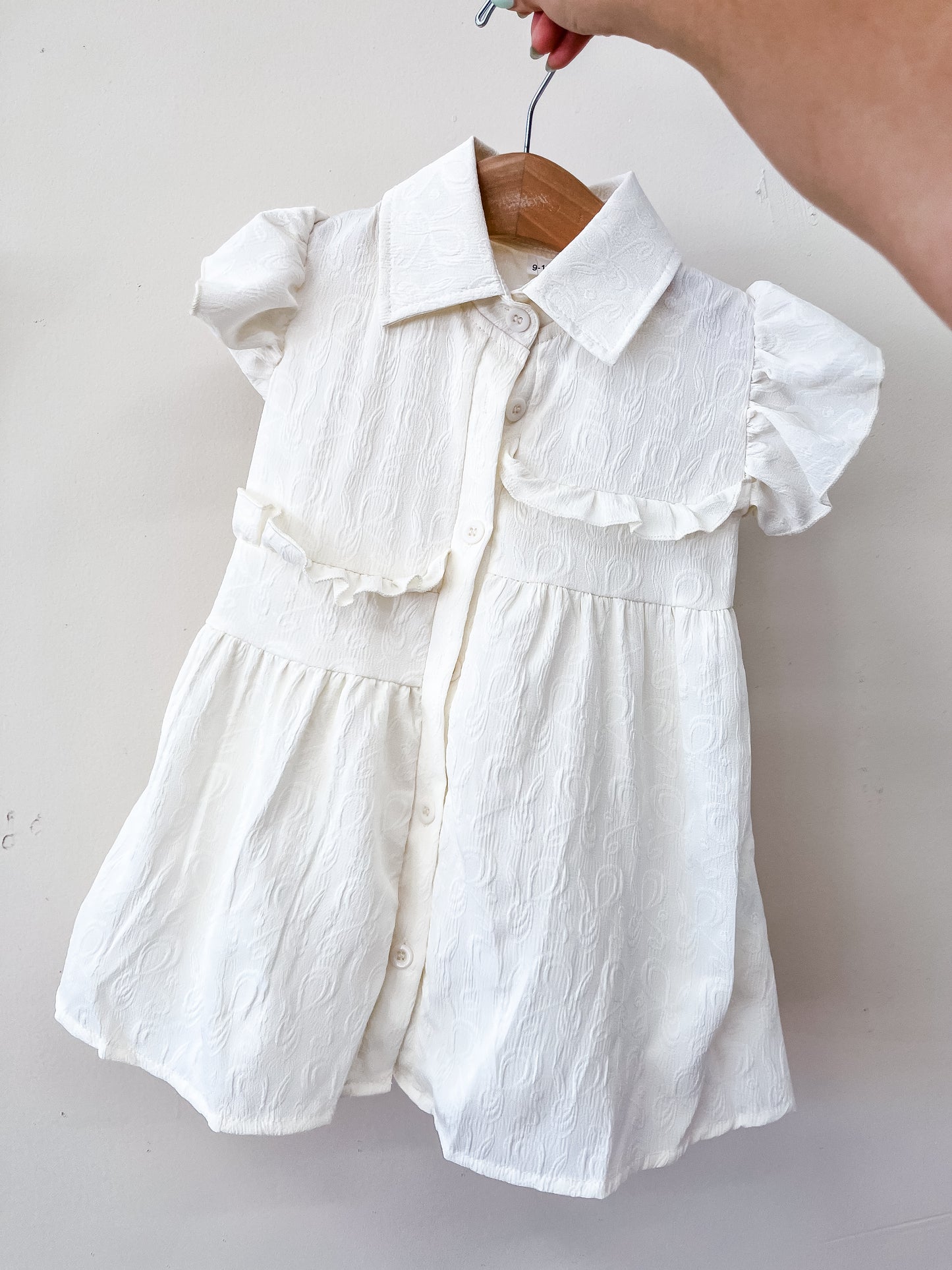 A hand holds a wooden hanger displaying the Willow Grace Dress by Tiny Tot Bundles, a white textured baby dress with short sleeves, a collar, chest ruffles, and a button-down front set against a plain light background.