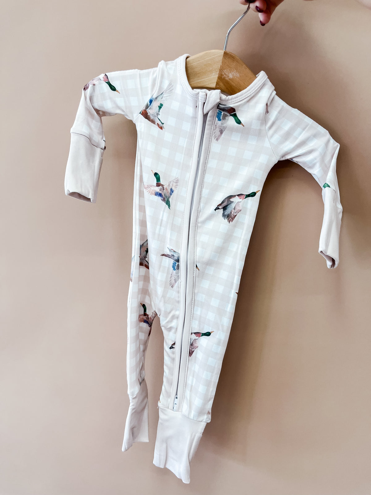 A hand holds the Tiny Tot Bundles Morning Flight Zippy on a hanger. This light-colored baby onesie features a subtle plaid pattern, charming bird illustrations, long sleeves, and a convenient front zipper.