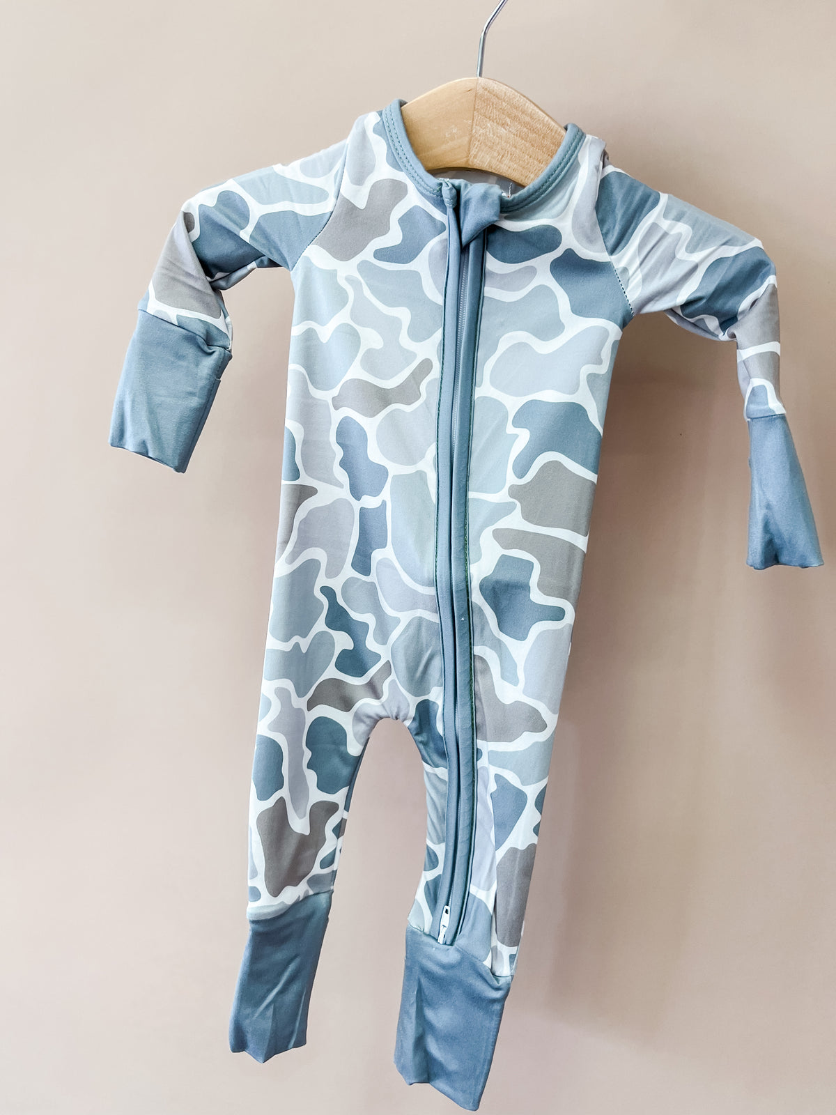 A Wild Country Zippy baby onesie by Tiny Tot Bundles, featuring a blue and gray camouflage pattern with long sleeves and a front zipper, hangs on a wooden hanger against a beige background.