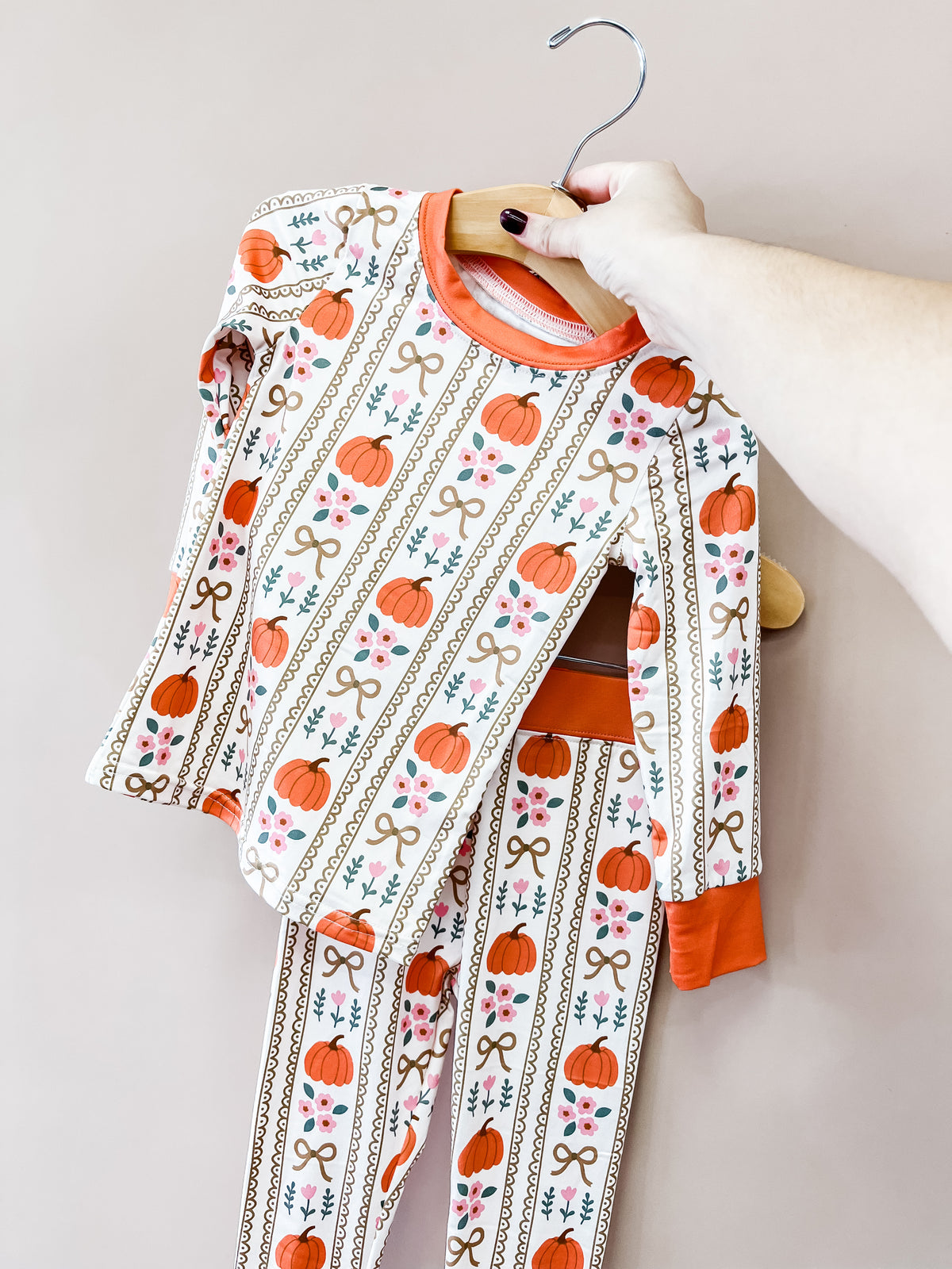 A hand holds an Autumn Bows Jammies Set, a two-piece pajama set featuring a pattern of pumpkins, bows, and flowers on a white background with orange trim.