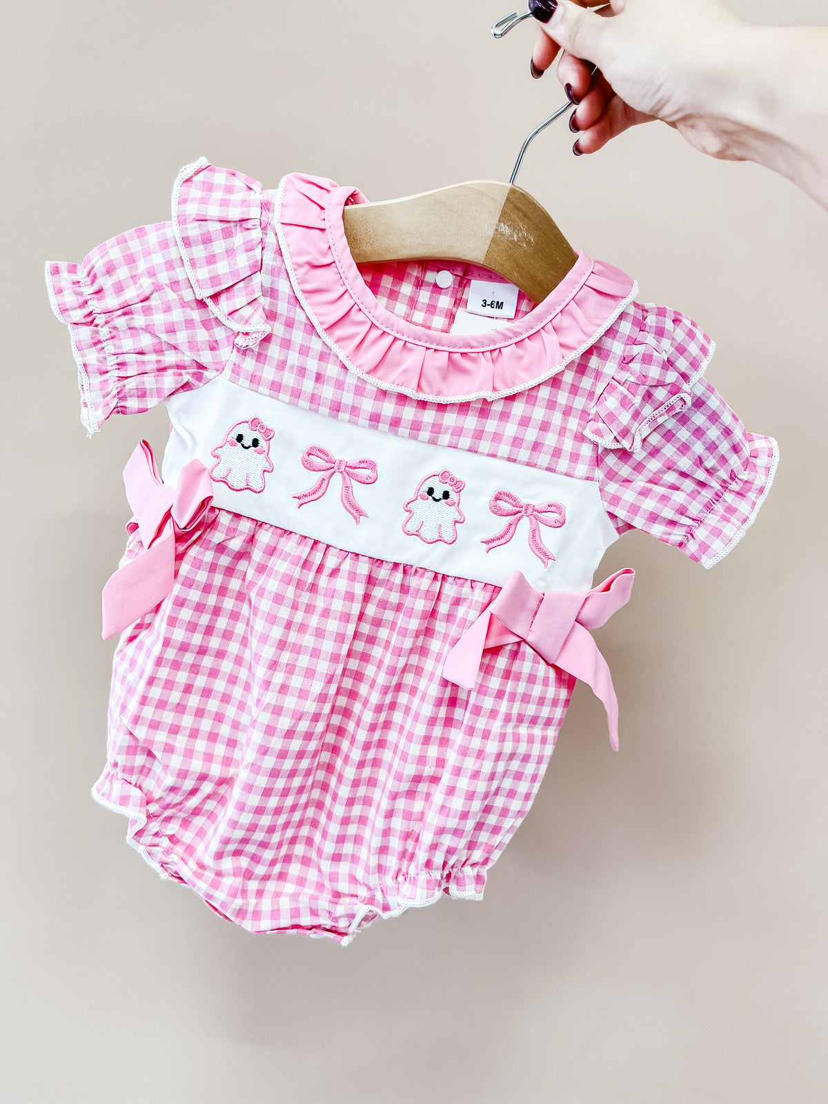 A hand holds the Pretty in Pink Ghost Bubble by Tiny Tot Bundles, a pink and white gingham romper with ruffled sleeves, bows, and embroidered cute animal faces on the chest.