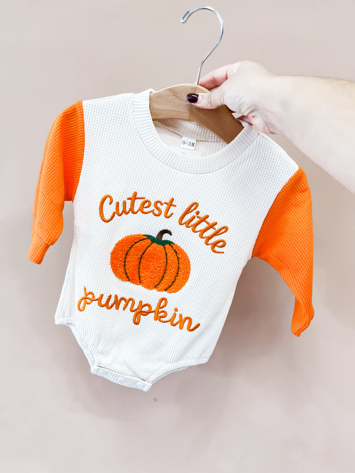 A hand holds the Tiny Tot Bundles Little Pumpkin Waffle Romper, sized 0-3M, on a wooden hanger. The off-white romper has orange sleeves, an embroidered pumpkin, and orange Cutest little pumpkin text.