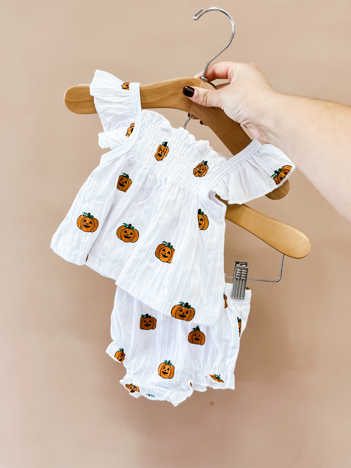A hand holds the Pumpkin Peekaboo Set by Tiny Tot Bundles: a white baby outfit with orange pumpkin prints, featuring a ruffled top and matching bloomers, against a tan background.
