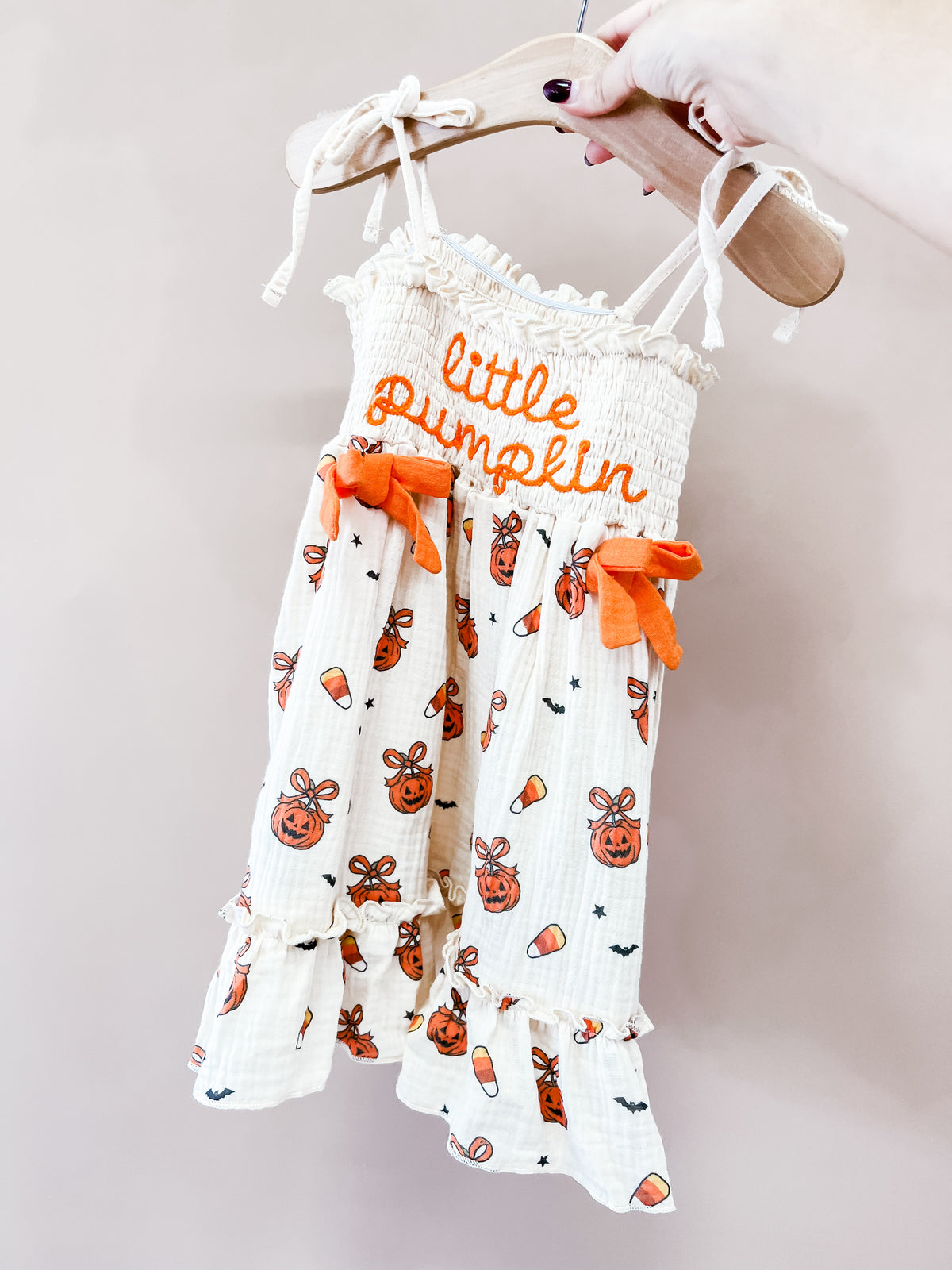 A hand holds the Tiny Tot Bundles Pumpkin Sweetheart Dress, a white toddler Halloween romper with orange pumpkin and candy corn prints, orange bows, and little pumpkin embroidered in orange on the chest.