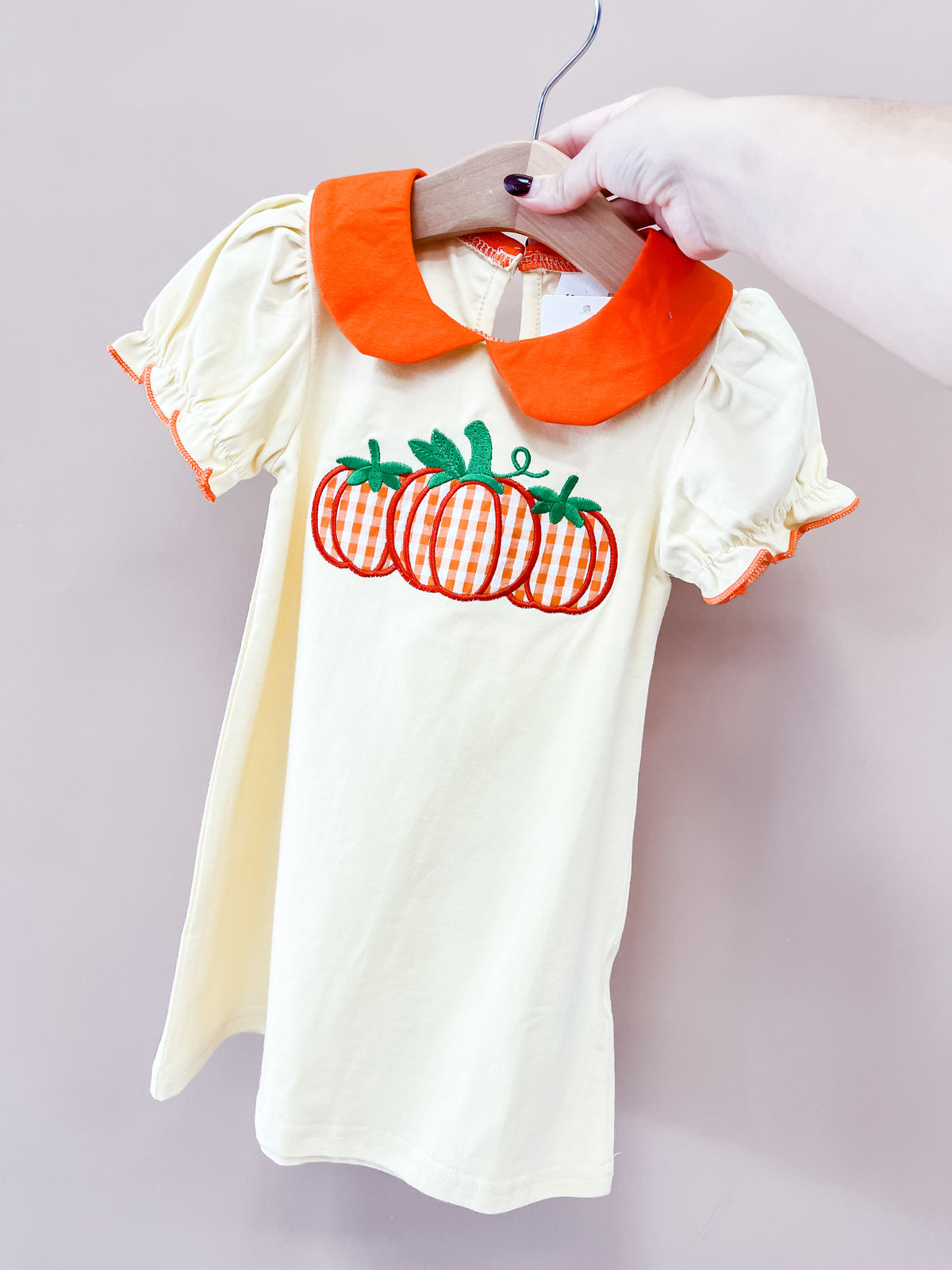 A hand holds the Tiny Tot Bundles Harvest Pumpkin Dress, a cream toddler dress with orange trim, puff sleeves, and three embroidered pumpkins with green leaves on the chest.